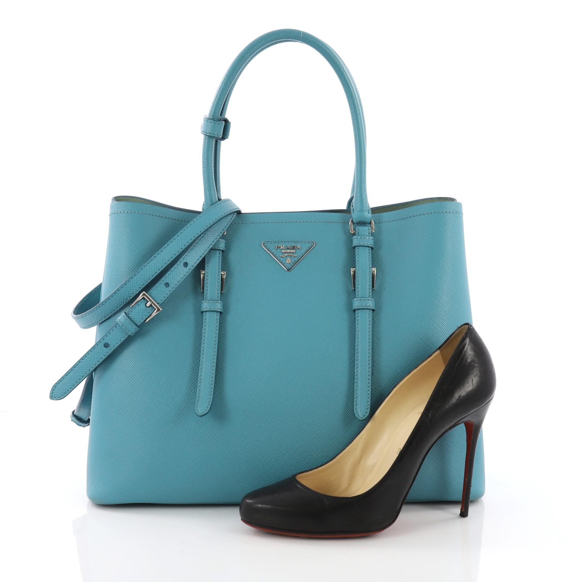 This Prada Cuir Covered Strap Double Tote Saffiano Leather Medium, crafted from blue saffiano leather, features dual rolled handles with extended belt accents, side snap buttons, and silver-tone hardware. It opens to a green leather interior with