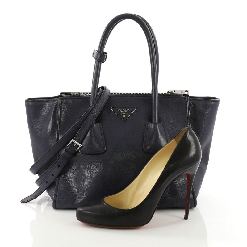 This Prada Twin Pocket Tote Glace Calf Small, crafted from blue glace calf leather, features tall dual rolled top handles, protective base studs, and silver-tone hardware. Its top snap closure opens to a black fabric interior with zip and slip