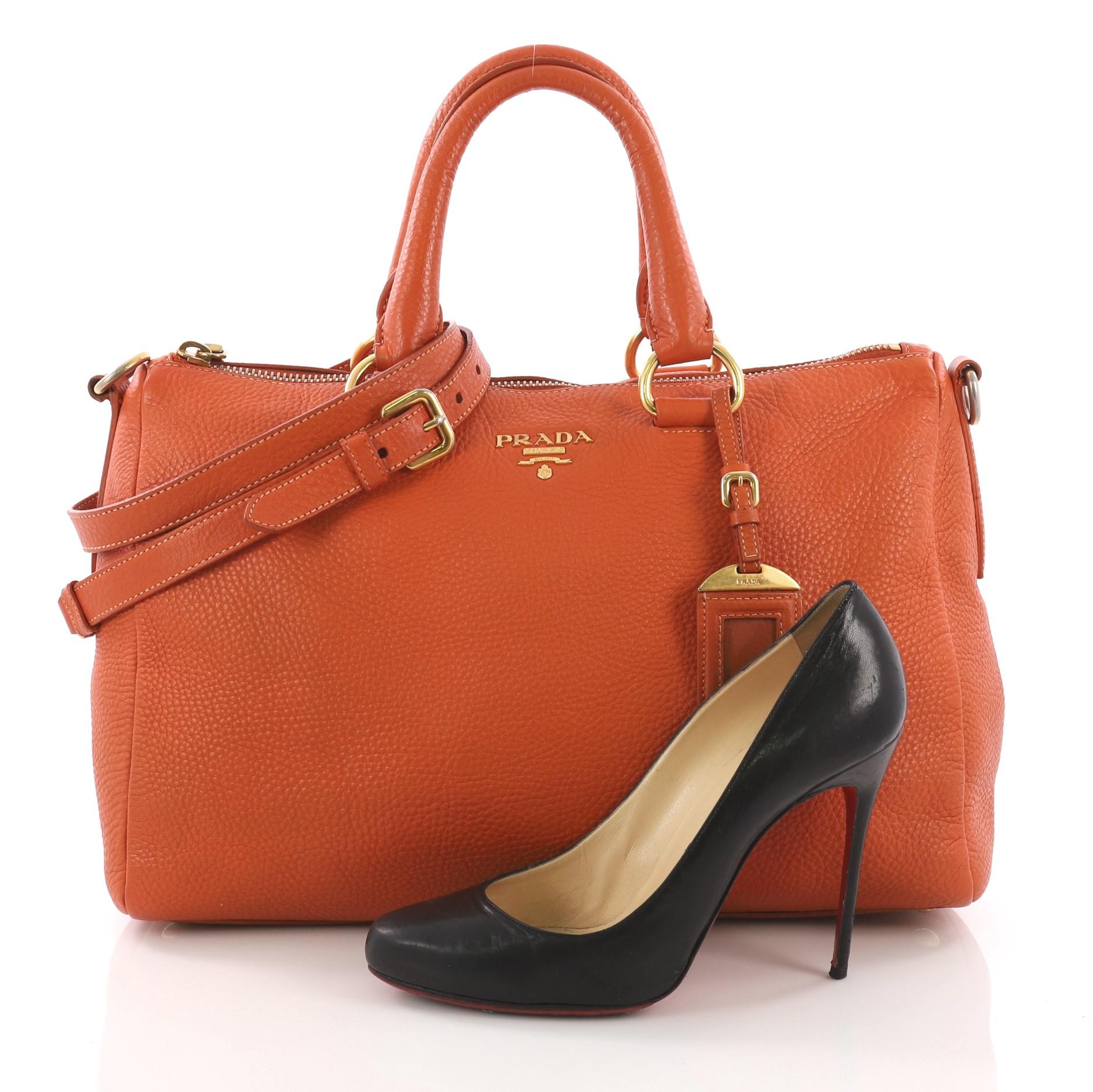 This Prada Convertible Boston Bag Vitello Daino Medium, crafted from orange vitello daino leather, features dual rolled leather handles, protective base studs, and gold-tone hardware. Its zip closure opens to an orange fabric interior with zip and