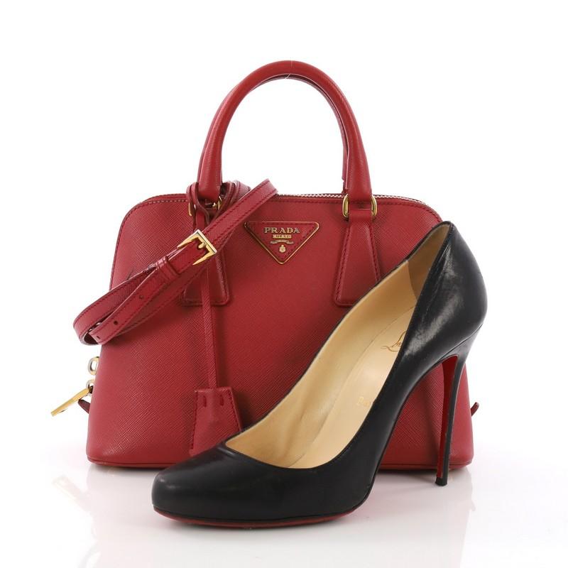 This Prada Promenade Handbag Saffiano Leather Small, crafted in red saffiano leather, features dual rolled leather handles, a detachable leather strap, protective base studs, and gold-tone hardware. Its zip closure opens to a red fabric interior
