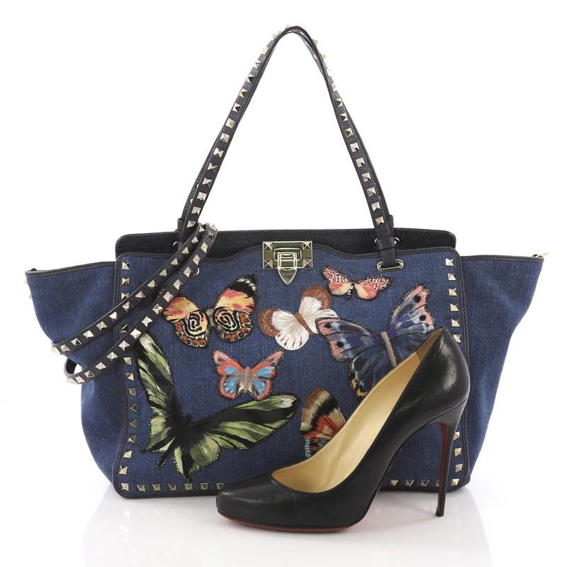 This Valentino Rockstud Tote Denim with Butterfly Applique Medium, crafted from blue denim with butterfly applique, features dual tall flat handles, pyramid stud trim details, and gold-tone hardware. Its clasp fastening opens to a blue denim