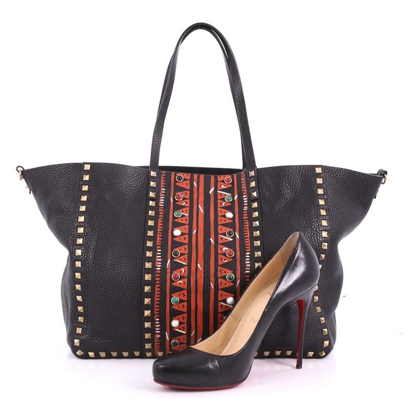 This Valentino Rolling Rockstud Convertible Tote Tribal Embellished Leather Medium, crafted from black embellished leather, features dual-flat leather handles, hand-painted tribal print design, cabochon and semi-precious stones, and matte gold-tone