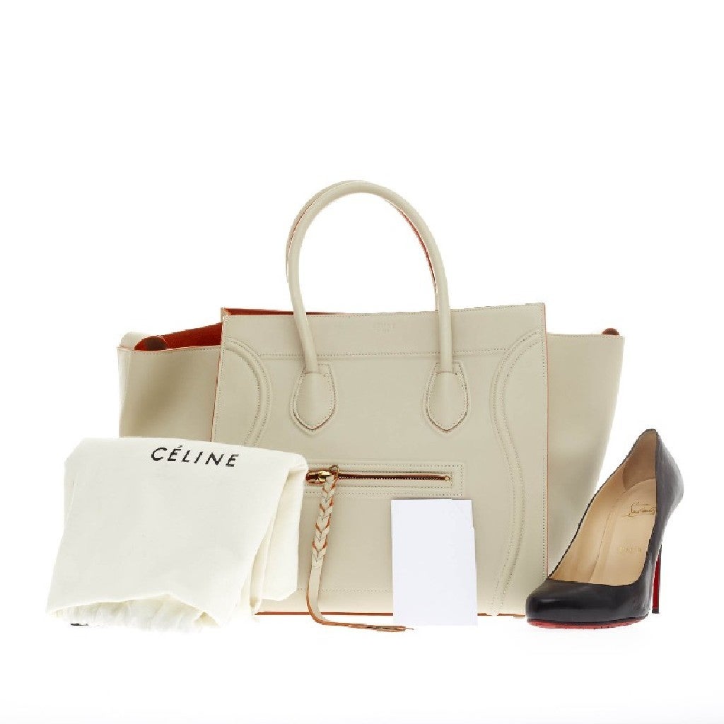 This authentic Celine Phantom Smooth Leather Medium is one of the most sought-after bags beloved by fashionistas. Crafted from sleek, off-white smooth leather with bright orange leather piping, this oversized minimalist tote features a braided