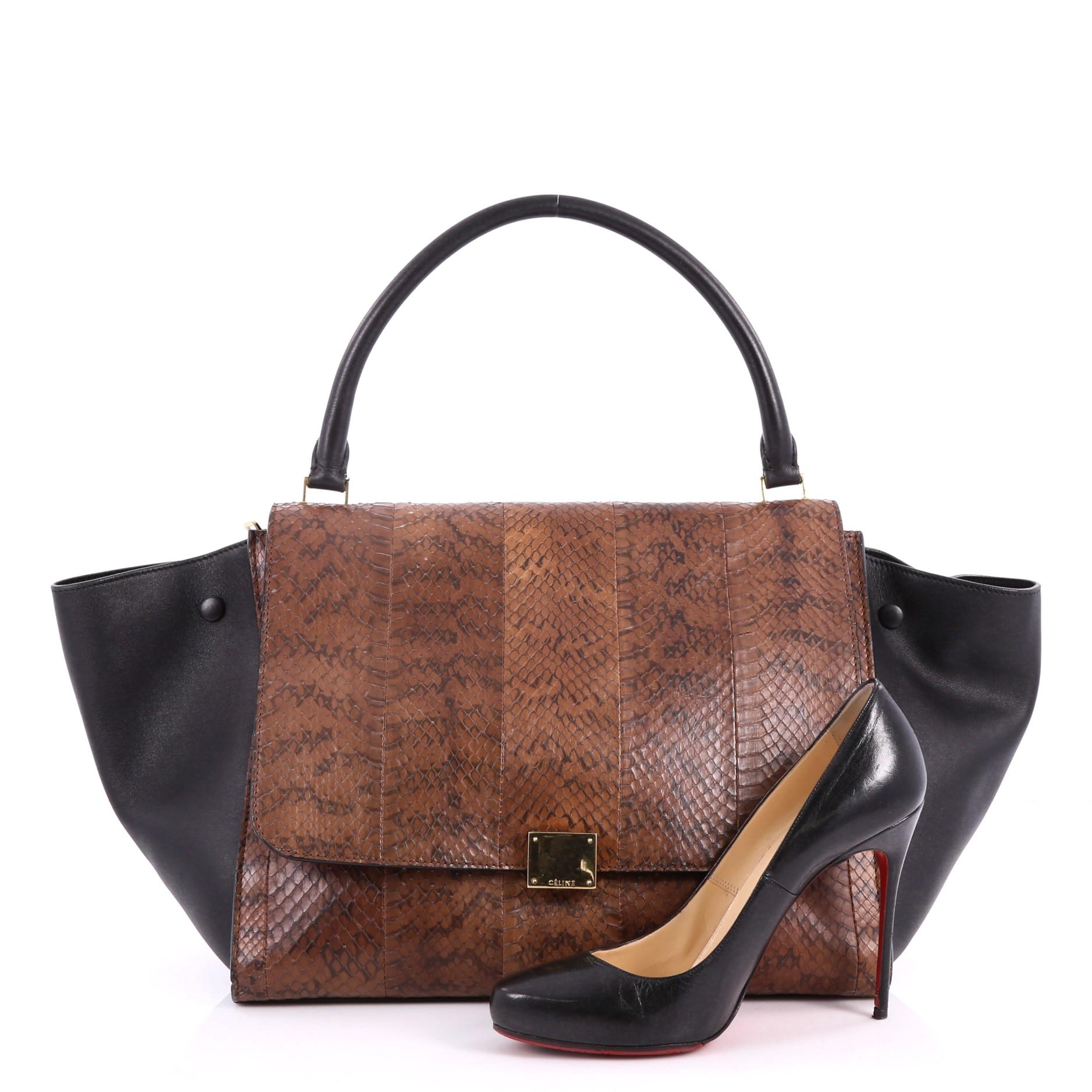 This authentic Celine Trapeze Handbag Python Large is a fashionista's dream. Crafted in genuine brown python skin with black leather wings, this stylish bag features exterior back zip pocket, top handle and gold-tone hardware accents. Its square