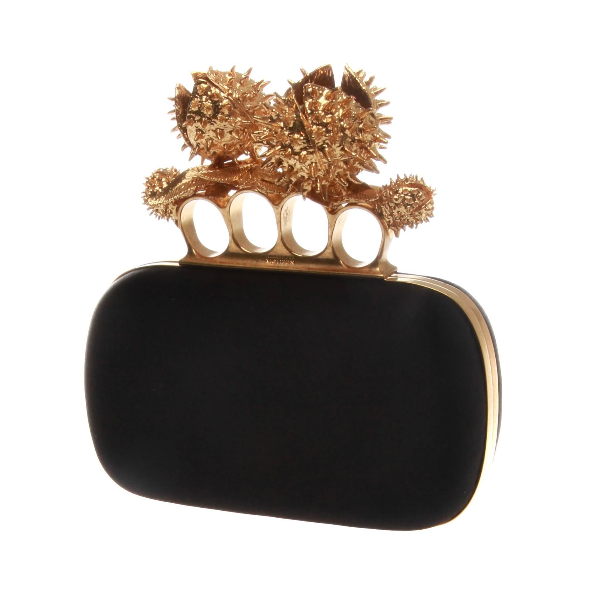 Stunning Alexander McQueen clutch of black satin starring a vibrant gold-tone metal elaboration. This masterfully crafted knuckle-duster style handle doubles as the fastening clasp and features open horse chestnuts with burgundy Swarovski stone