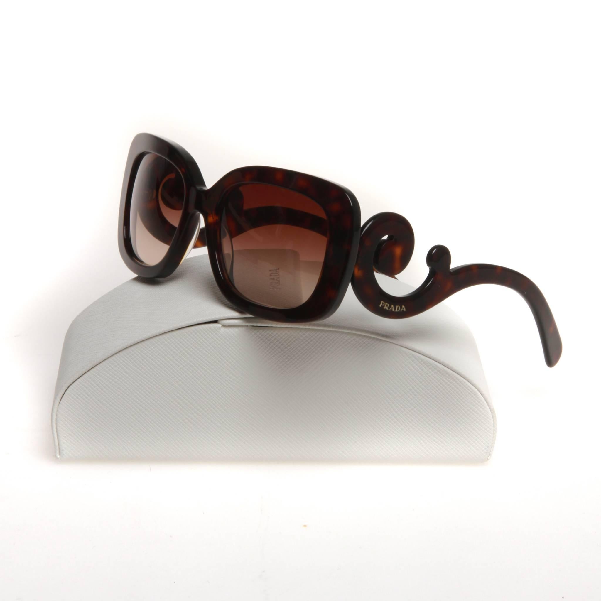 Brown tortoiseshell acetate Prada sunglasses with Baroque accents at temples featuring gold-tone logo and gradient lenses. Includes case, cloth and box.