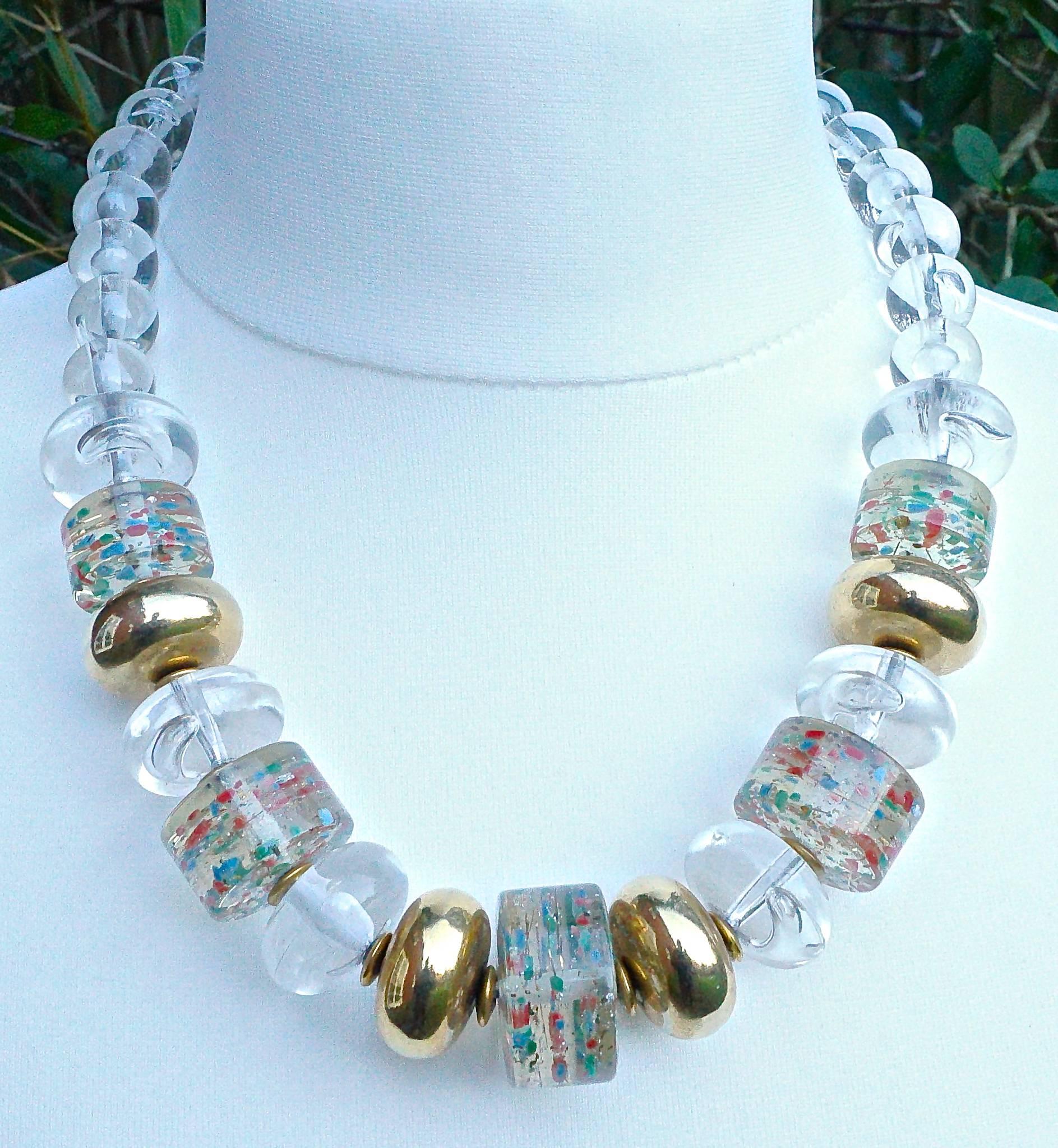 Large plastic bead necklace with clear and golden beads, and featuring five disc shaped beads with blue, green, red and silver confetti. Most of the clear beads have clear bubbles inside. There is a length of smaller round clear beads leading to the