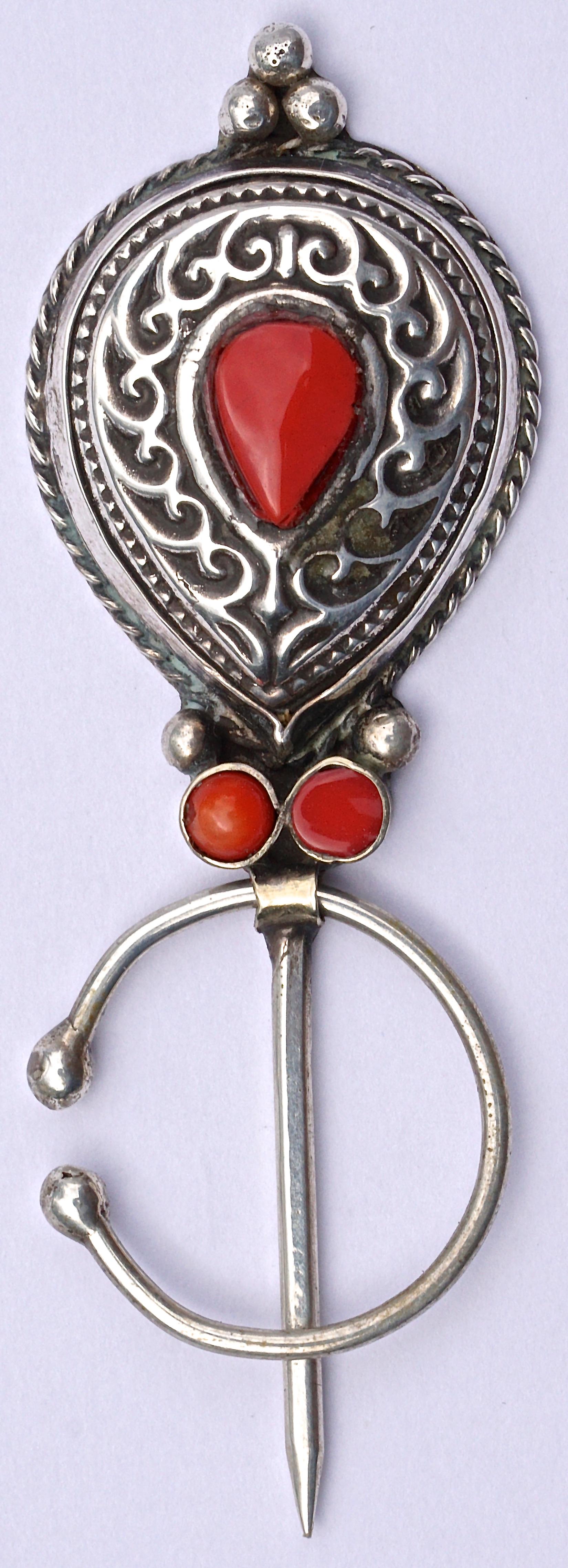 Moroccan silver penannular brooch with glass coral stones, and featuring a swirl design with rope twist edging. It is  handmade by Berbers, an ethnic people indigenous mainly to Morocco and Algeria. The brooch pins to the garment and is then locked