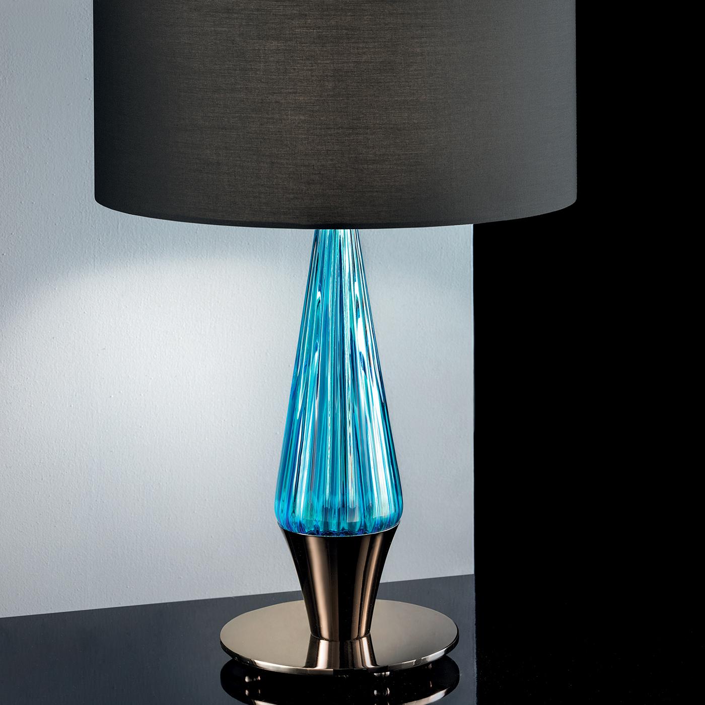 This lamp is characterized by an innovative conical shaft with a rectangular section and a short cylindrical lampshade in gray polycotton fabric and pvc. It stands out thanks to its aquamarine details that give a splash of freshness to its design.