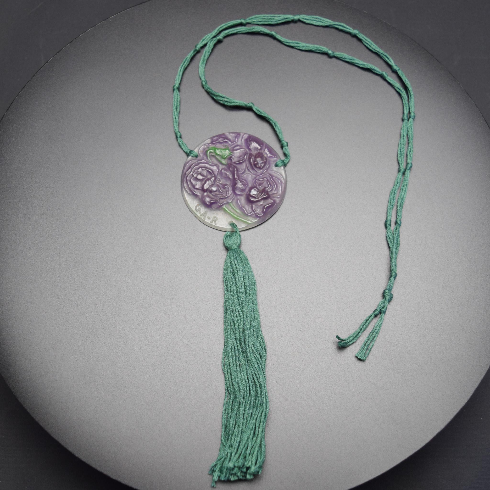 Argy Rousseau Pate de verre Parma violets pendant C1921
Circular flat moulded pendant with relief floral decoration.
Signed G.A.R  
Strung with a knotted green neck cord and tassel.

Reference -   See page 186 G Argy Rousseau glassware as art
Janine