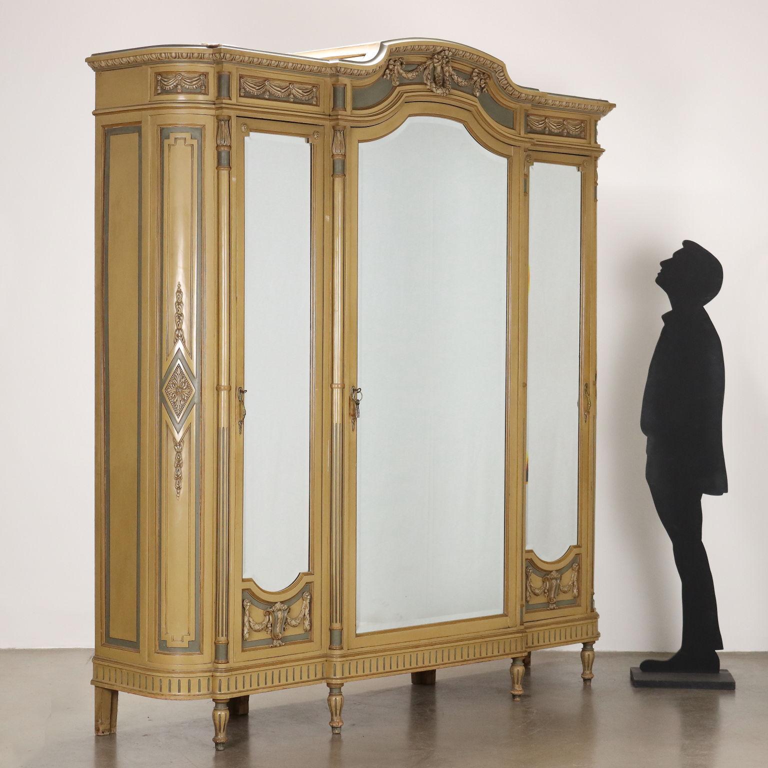 Armadio in stile Neoclassico in faggio laccato, produzione Fabbrica di Mobili “Luigi Gatti” Torino. Fronte a tre ante con specchi bisellati, frapposti a colonne con sgorbiature, intagli con festoni e drappeggi nella fascia superiore e nei pannelli