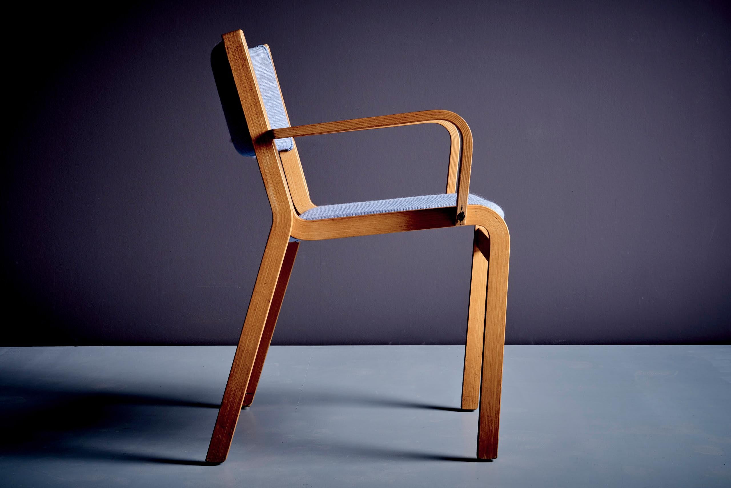 Este sillón, diseñado por Rud Thygesen y Johnny Sørensen y fabricado por Magnus Olesen en Dinamarca, muestra la refinada artesanía moderna danesa. Con un armazón de madera curvada de roble en capas y tapizada en tela de lana azul texturizada, la