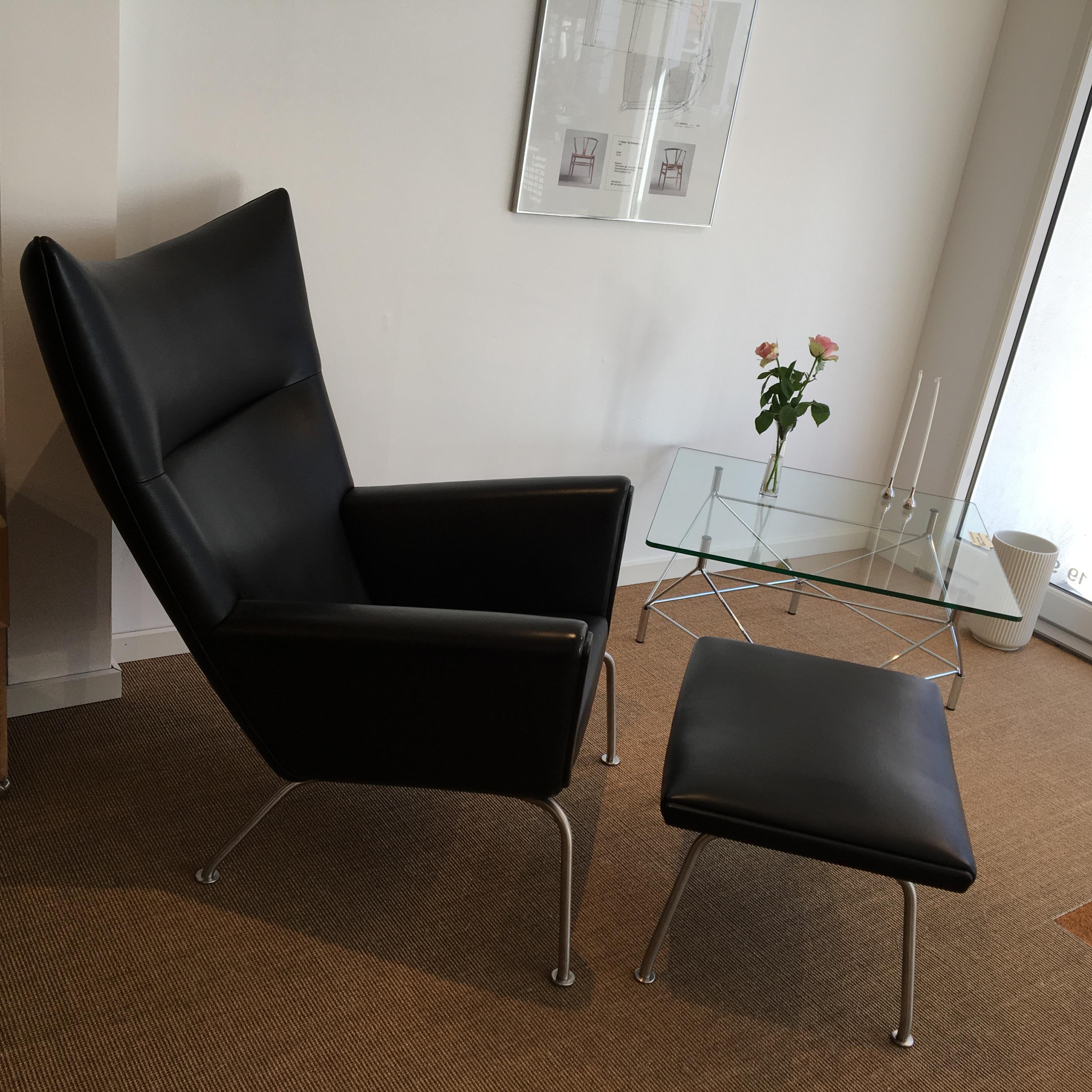Armchair, 'Wing Chair' with footstool CH445 and CH446 upholstered in black leather (Thor) and stainless steel legs.
Design: Hans J. Wegner. Designed in 1960.
Dimensions: D 90, B 90, H 105 cm, SH 39 cm.
Dimensions of stool: 63 x 50 cm SH 38 cm.
 