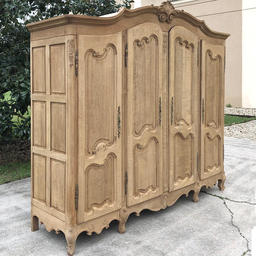 Antique Country French stripped four-door armoire was crafted from solid oak, and features a gracefully arched crown atop the doors that possess elegantly scrolled frameworks. Each door opens to reveal a cavernous interior. Scrolled and carved apron