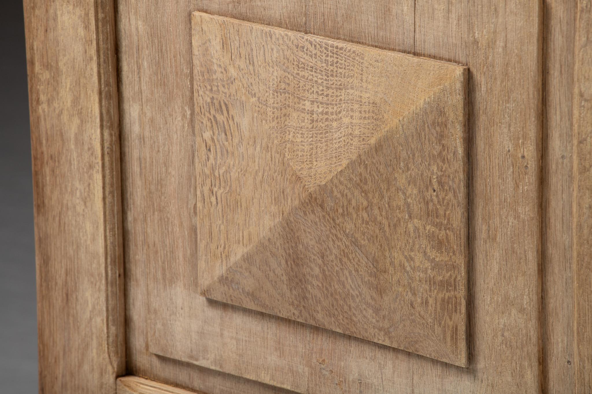 Standing with monastic presence, this tall armoire in natural oak bears a façade of geometric rigor and quiet monumentality. Six raised diamond-point panels animate the surface with a rhythm of shadow and light, their pyramidal forms subtly