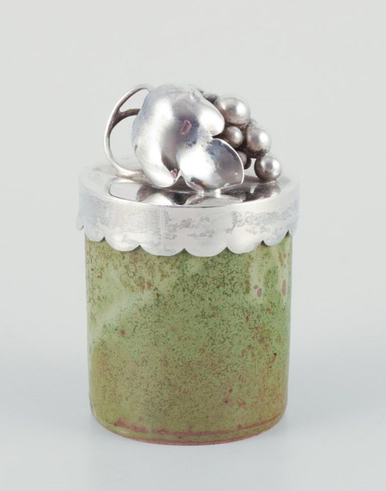 Arne Bang/Aage Weimar, lidded jar. Ceramic jar with green-toned glaze, sterling silver lid. Adorned with a cluster of grapes.
Model number 157.
Mid-20th century.
Silver lid stamped AAW 925.
Jar signed.
In perfect condition.
Dimensions: Height 8.0 cm