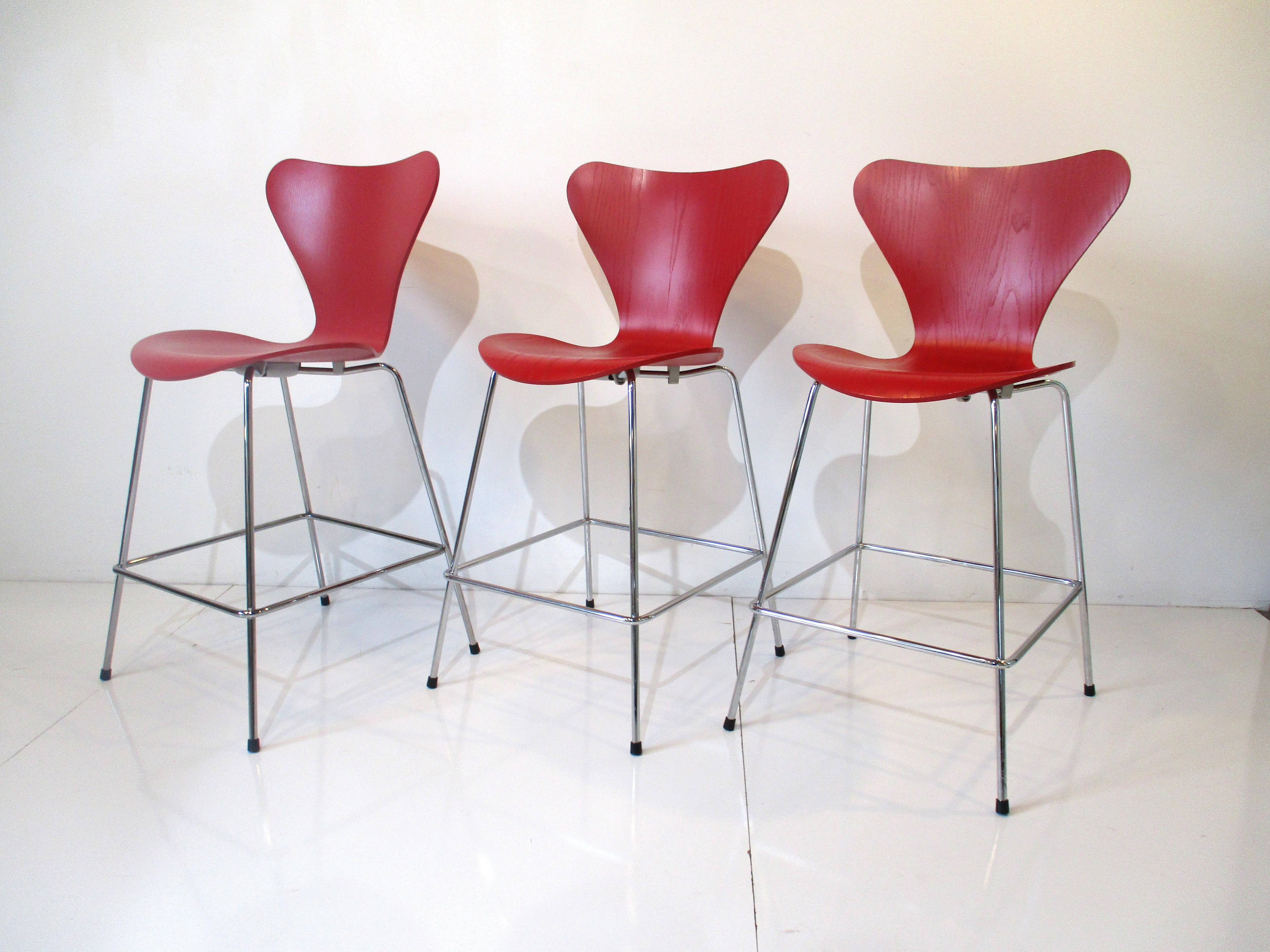 A set of three matching red molded plywood seats on chromed metal framed bar / counter stools with foot rests . Designed by Arne Jacobsen and manufactured by Fritz Hansen made in Denmark , the chairs are not knock offs and retain the original Hansen