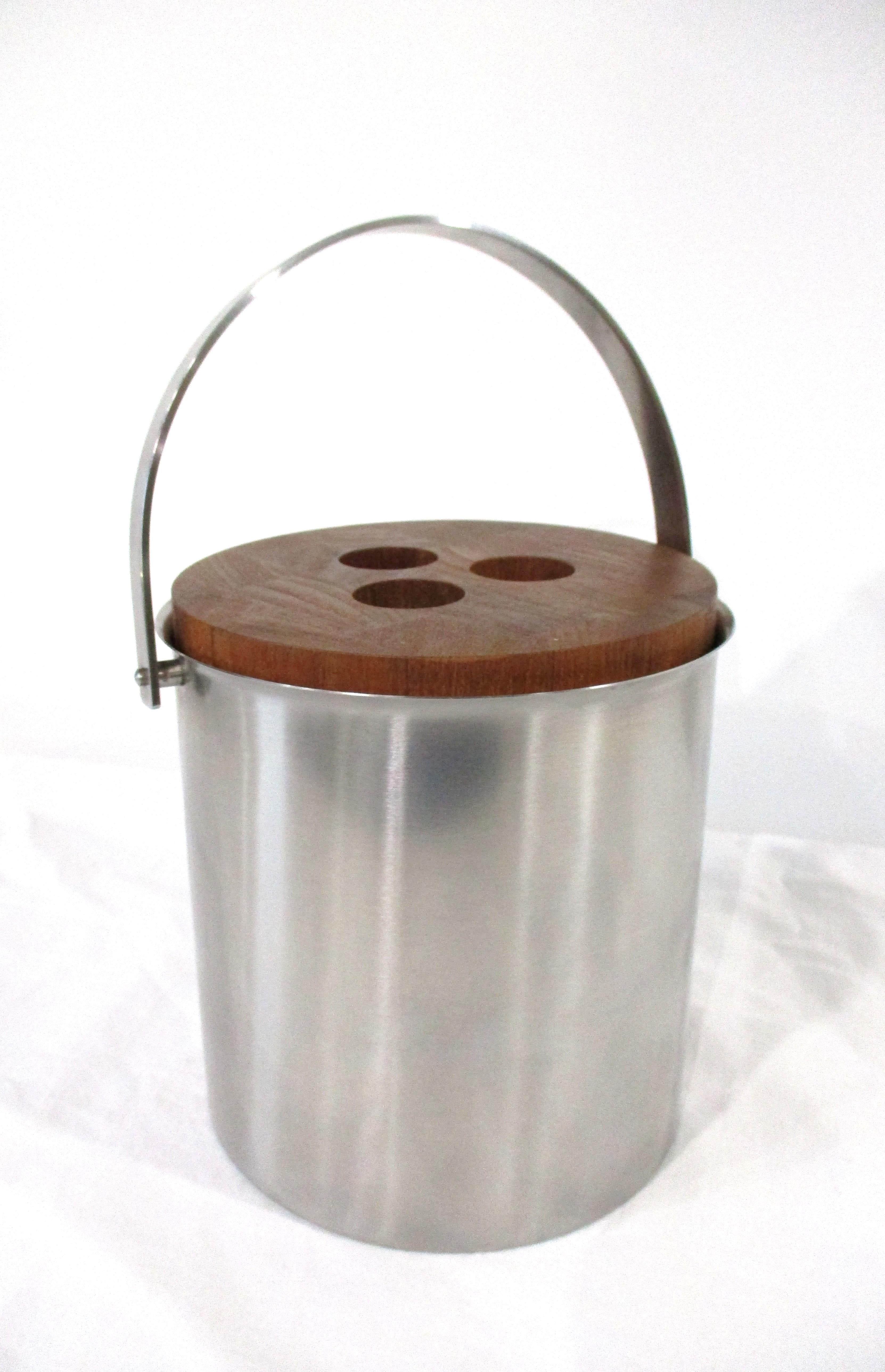 A stainless-steel ice bucket with a teak wood lid having three finger holes and folding handle. A black plastic insert inside for easy cleaning a very well-crafted quality example of design work by Arne Jacobsen made in Denmark by Stelton, perfect