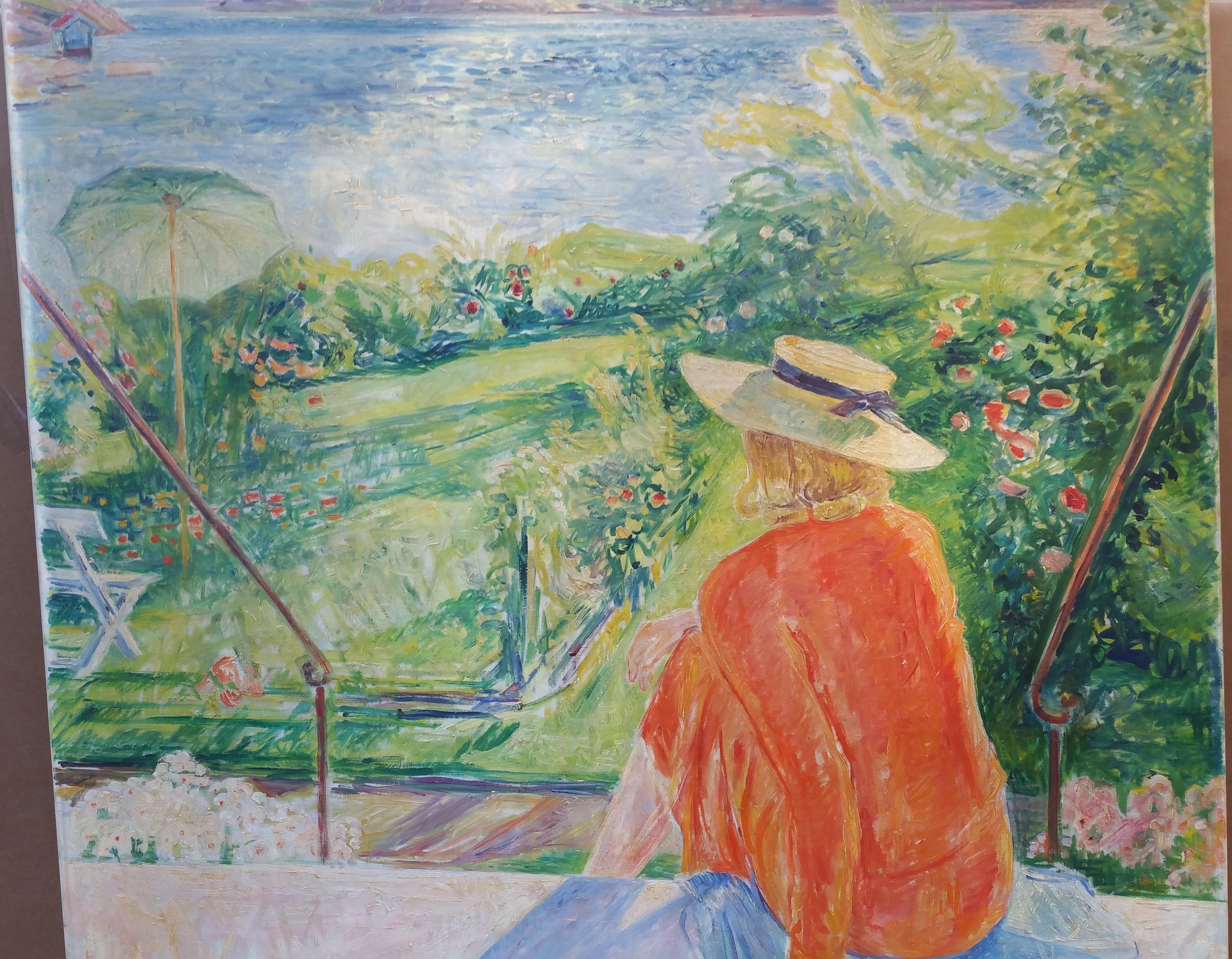 Signed lower left.

A blond woman clad in a red blouse and skirt sits atop a blue blanket at the top of a set of stairs leading to a garden at the edge of a large body of water, with wooded hills in the distant background. Her back is turned from