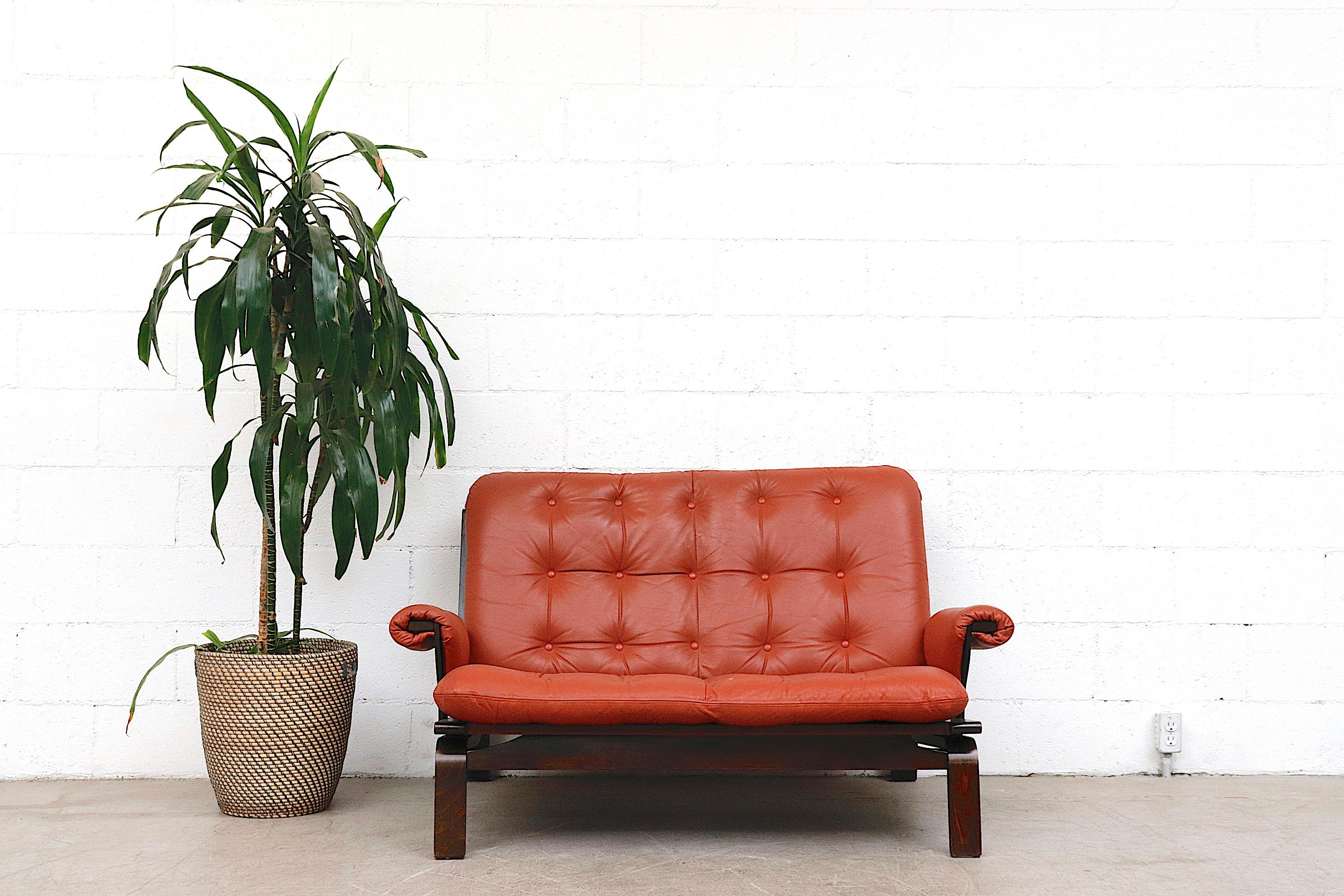 Arne Norell style coral leather loveseat with lightly refinished bentwood frame and padded leather arm rests. Canvas sling support in original condition with visible fading. Good overall condition. Wear is consistent with its age and usage.