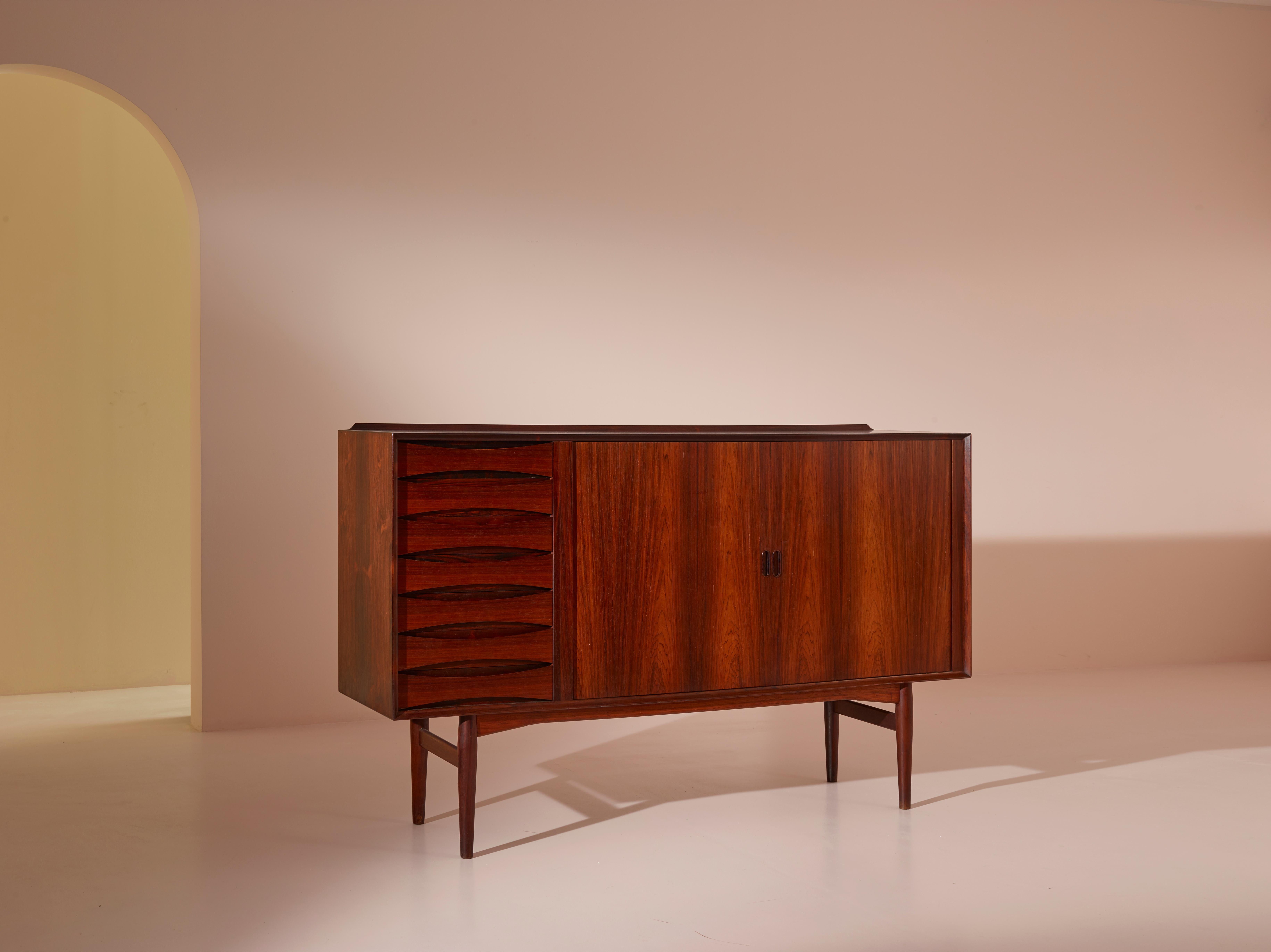 The OS63 highboard, designed by Arne Vodder for Sibast Mobler in 1958, is a beautiful and functional storage piece made of rosewood. With seven drawers on the left and tambour doors on the right, this credenza provides ample storage space while