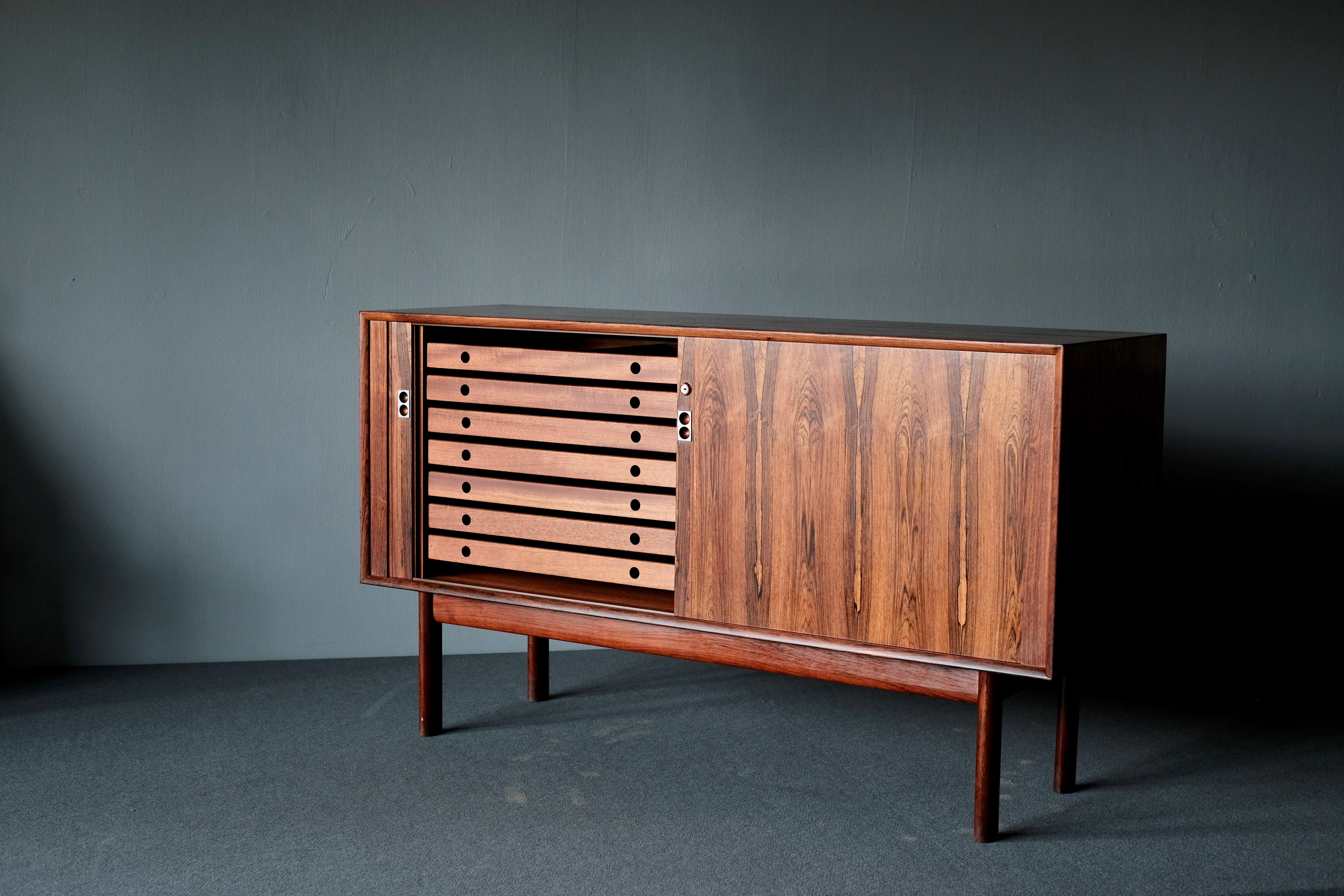 Sideboard by Arne Vodder for Sibast Mobler. It is in wonderfully patinated rosewood. The front has two ingenious tambour doors, enclosing trays and shelves. The handles are in steel. This Vodder sideboard with its modernism and perfect proportions