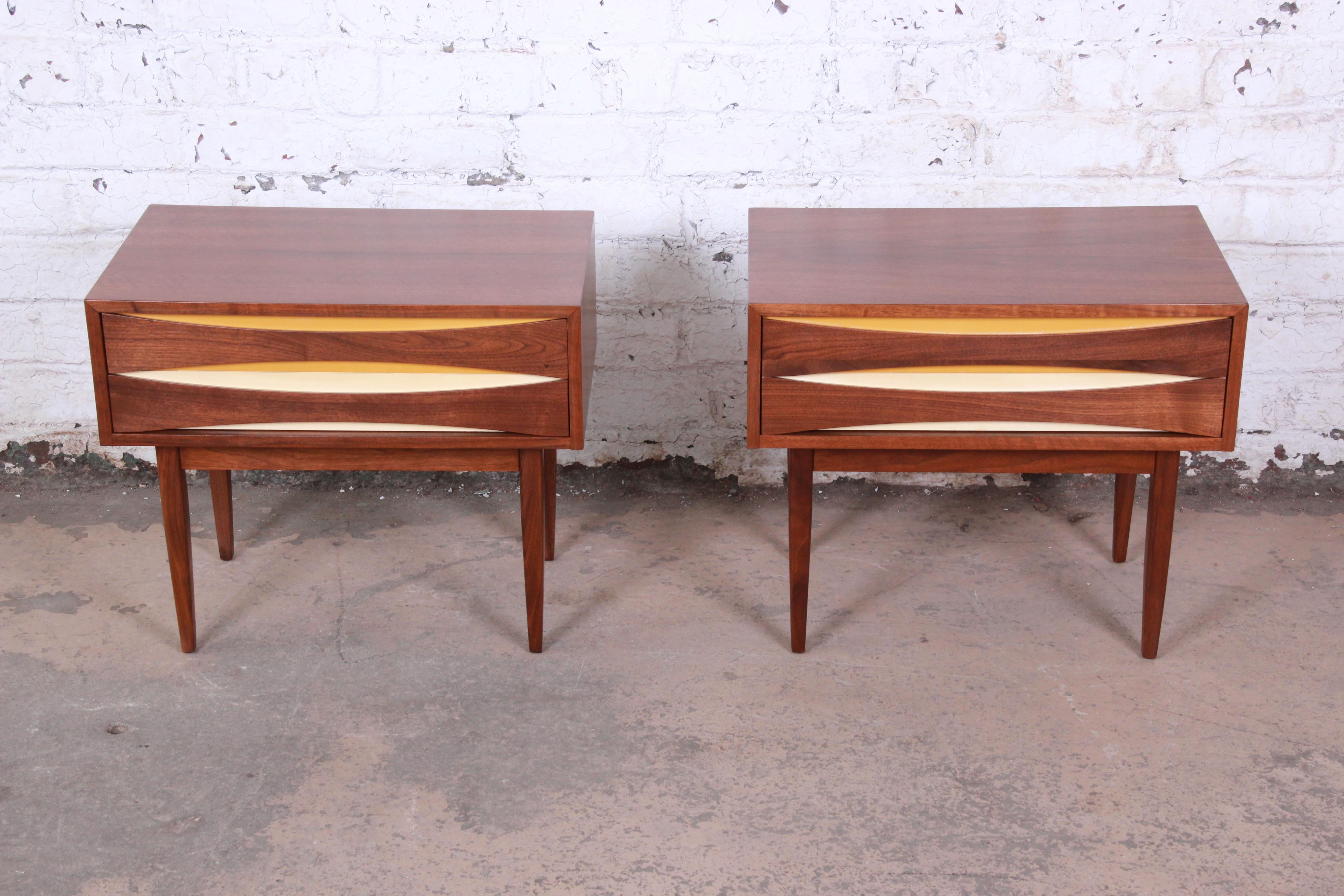An exceptional pair of Arne Vodder style Mid-Century Modern walnut nightstands by West Michigan Furniture. The nightstands have a beautiful walnut wood grain with sculpted pulls with a goldenrod and white backdrop on the drawers. They offer good