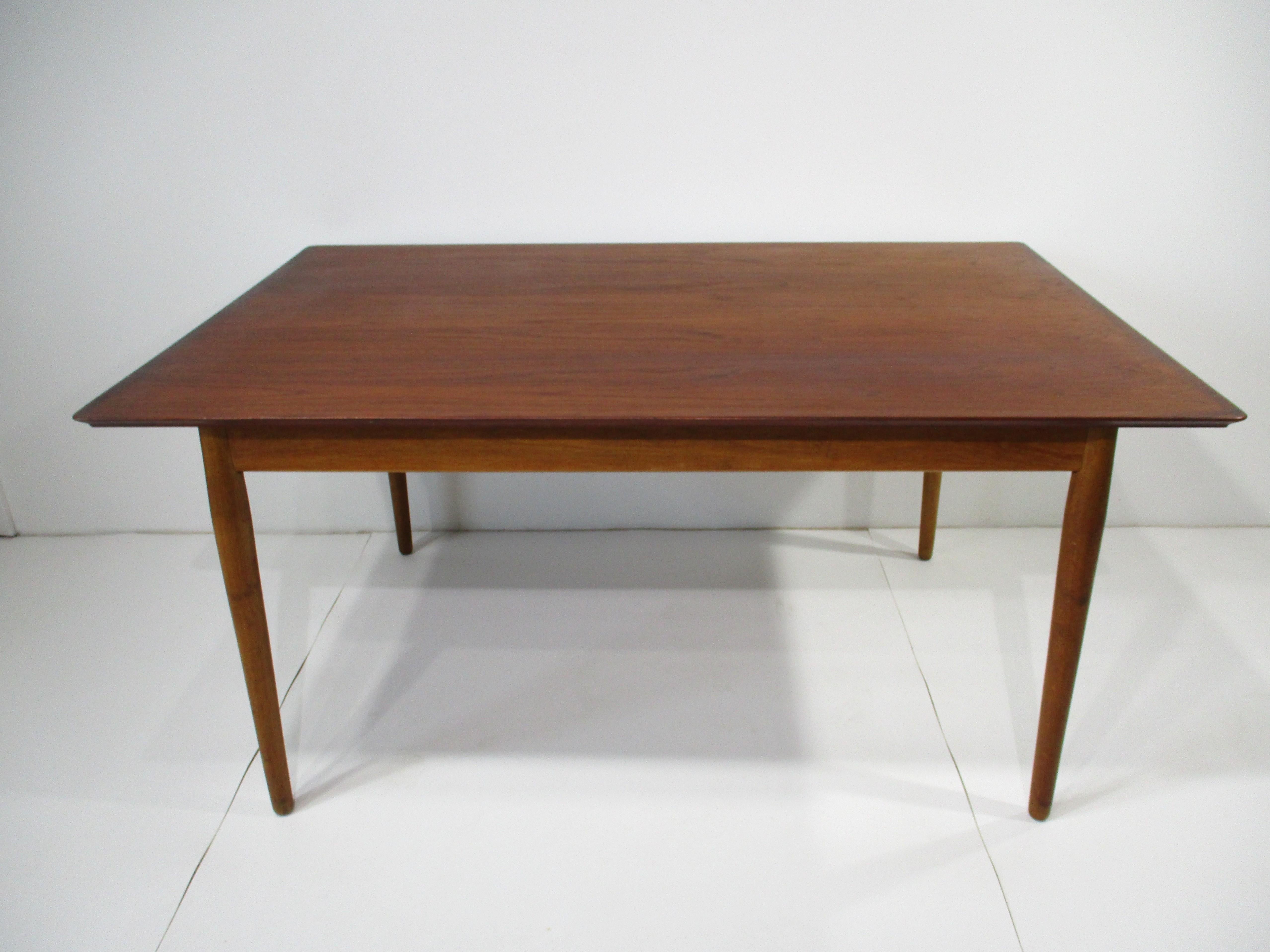 A medium dark teak wood expandable dining table with two slip in end leaves all sitting on tapered legs . A trim of darker teak surrounds the top boarder edge of the table and shows the superb quality and craftsmanship of early cabinet making by the