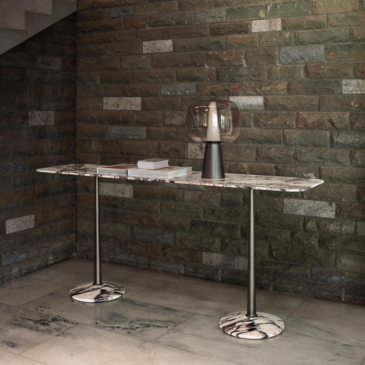 The Arnold Console features a refined combination of geometric forms—a tapered column, a flat surface, and a truncated conical base—crafted with precise proportions. The design highlights an elegant brass column that connects the slim, rounded-edge