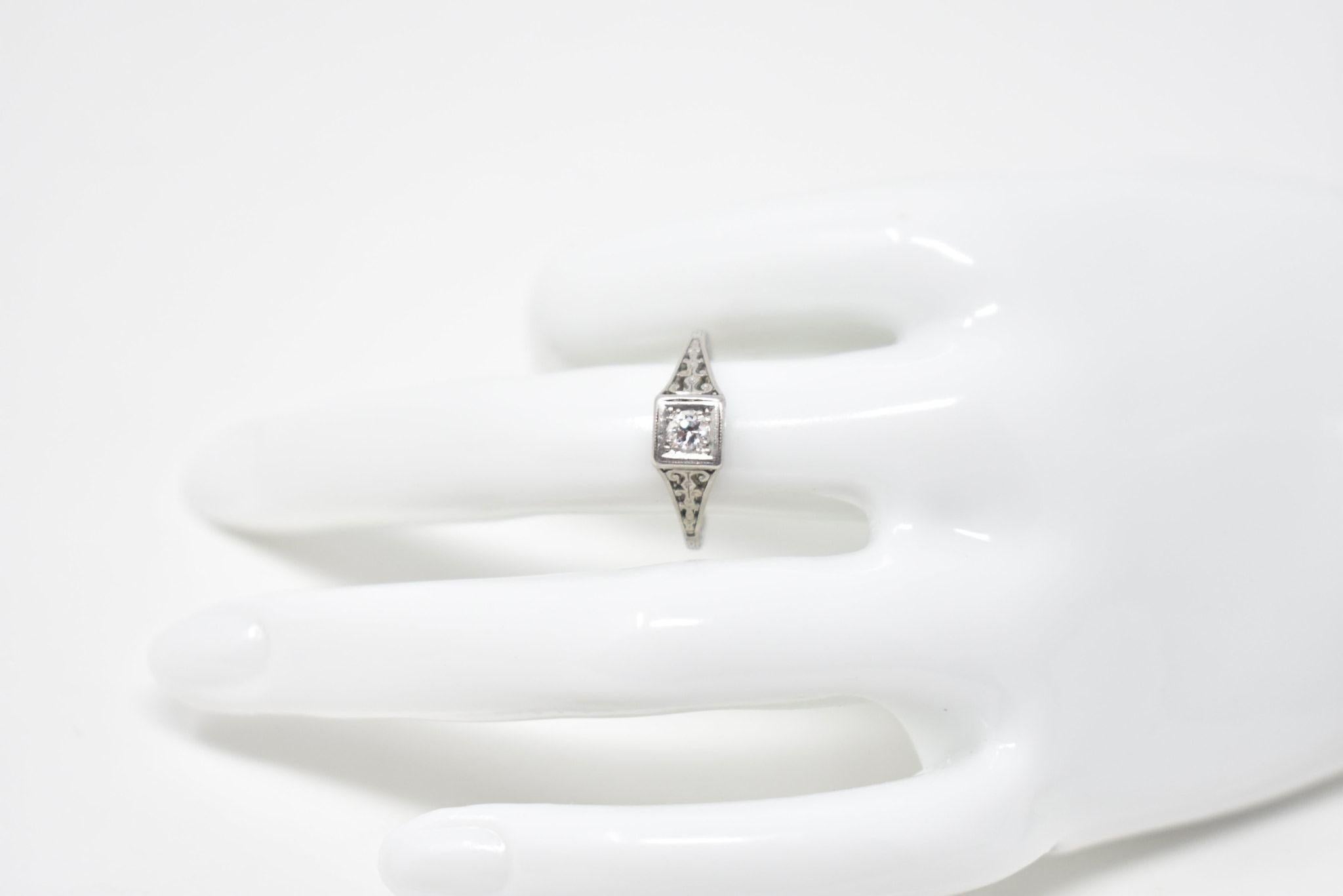 This is a lovely diamond engagement ring that has delicately scrolled filigree work on the shoulders. It is set in rich platinum and centering an old European cut diamond weighing approximately 0.15 carats, G color and VS clarity. The top of the