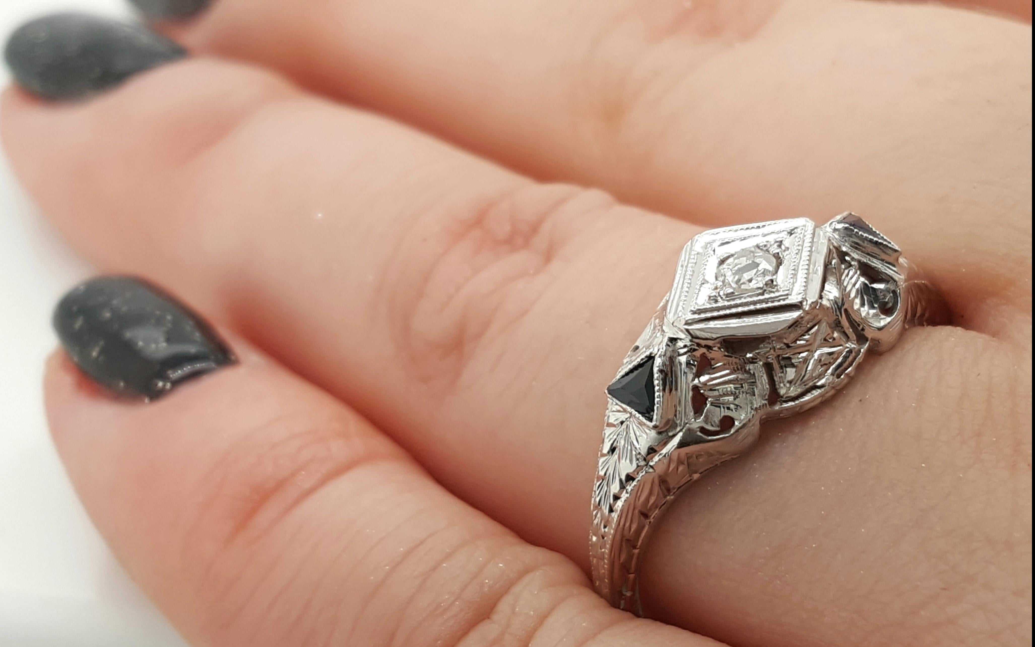 This lovely example of an Art Deco ring is crafted of stunning 18K white gold. Intricate filigree designs surround one center diamond on this vintage beauty. Two sapphire pyramids add a pop of color that adds to this already beautiful piece. This is