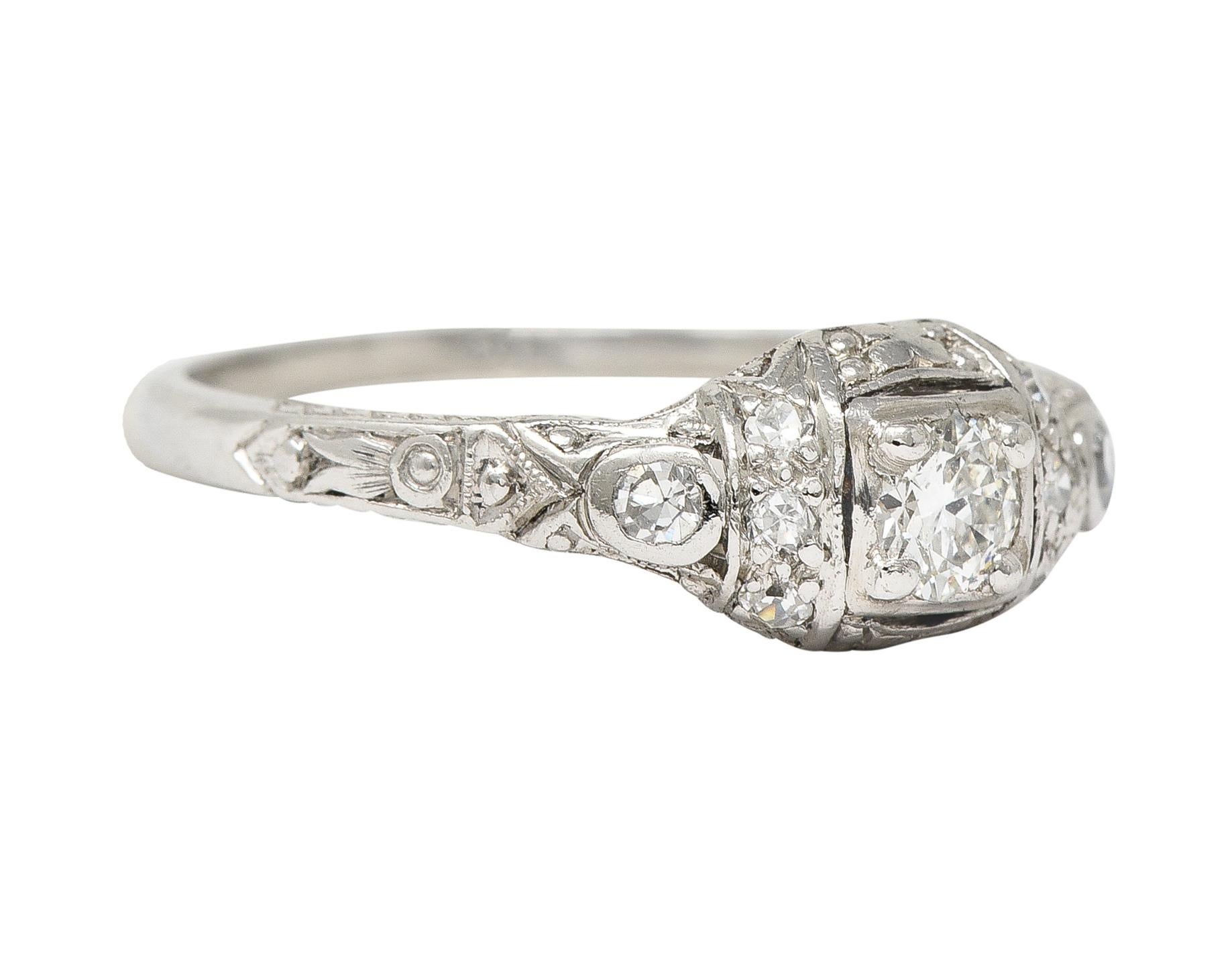 Centering a transitional cut diamond weighing approximately 0.15 carat - I/J color and SI clarity
Bead set in a square form head with a pierced and engraved ornate geometric surround
Flanked by channel and bezel set single cut diamonds weighing