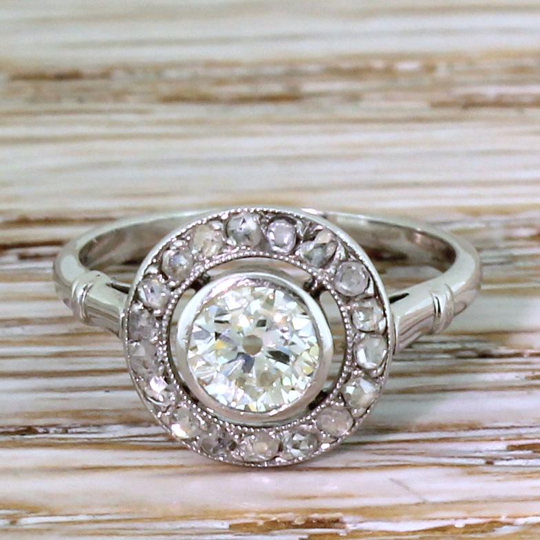 A vintage target cluster ring of class and beauty. The old European cut diamond in the centre is rubover set, and floats within a halo of eighteen rose cut diamonds set in milgrained white gold. The cluster sits nice and low to the finger on a slim
