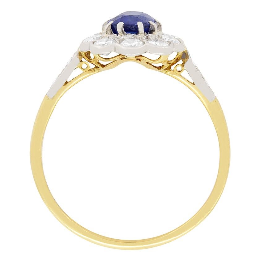 A stunning blue sapphire is encircled by transitional cut diamonds in this Art Deco ring. The 0.80 carat sapphire is oval shaped, and is a mesmerising blue colour. Ten diamonds surround the sapphire in a daisy style halo, and extend to cover the