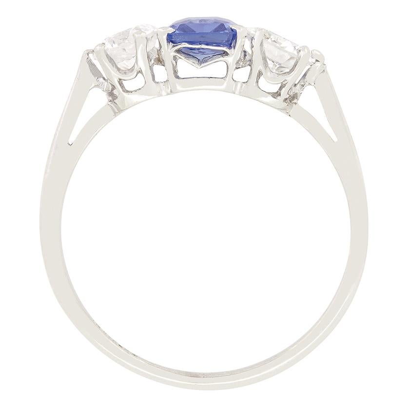 A gorgeous cornflower blue Sapphire sits between two transitional cut diamonds in this Art Deco trilogy ring. The natural 0.80 carat sapphire is an emerald cut stone and is claw set into 18 carat white gold. The flanking diamonds are 0.40 carat a