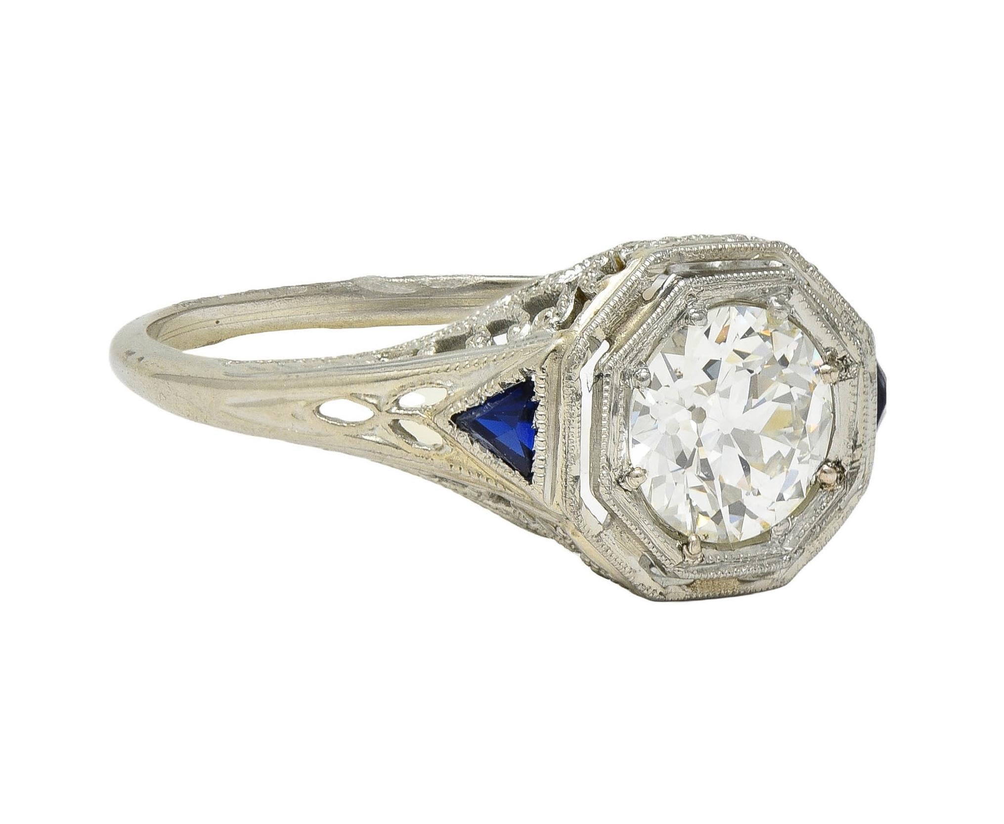 Centering an old European cut diamond weighing approximately 0.91 carat - H color with VS2 clarity
Bead set in an octagonal form head with a pierced milgrain surround 
Flanked by triangular calibré cut synthetic sapphires bezel set at