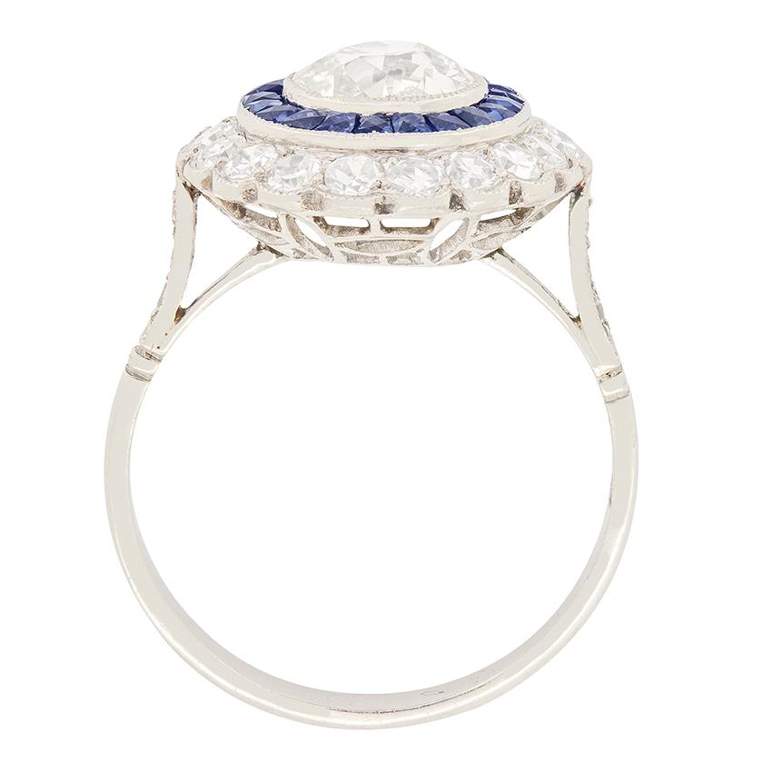 A wonderful 1.10 carat old cut diamond is enveloped in a ring of sapphires in this Art Deco target ring. The central diamond is rub over set with milgrain detailing across the setting. The surrounding circle of sapphires totals to 0.63 carat of
