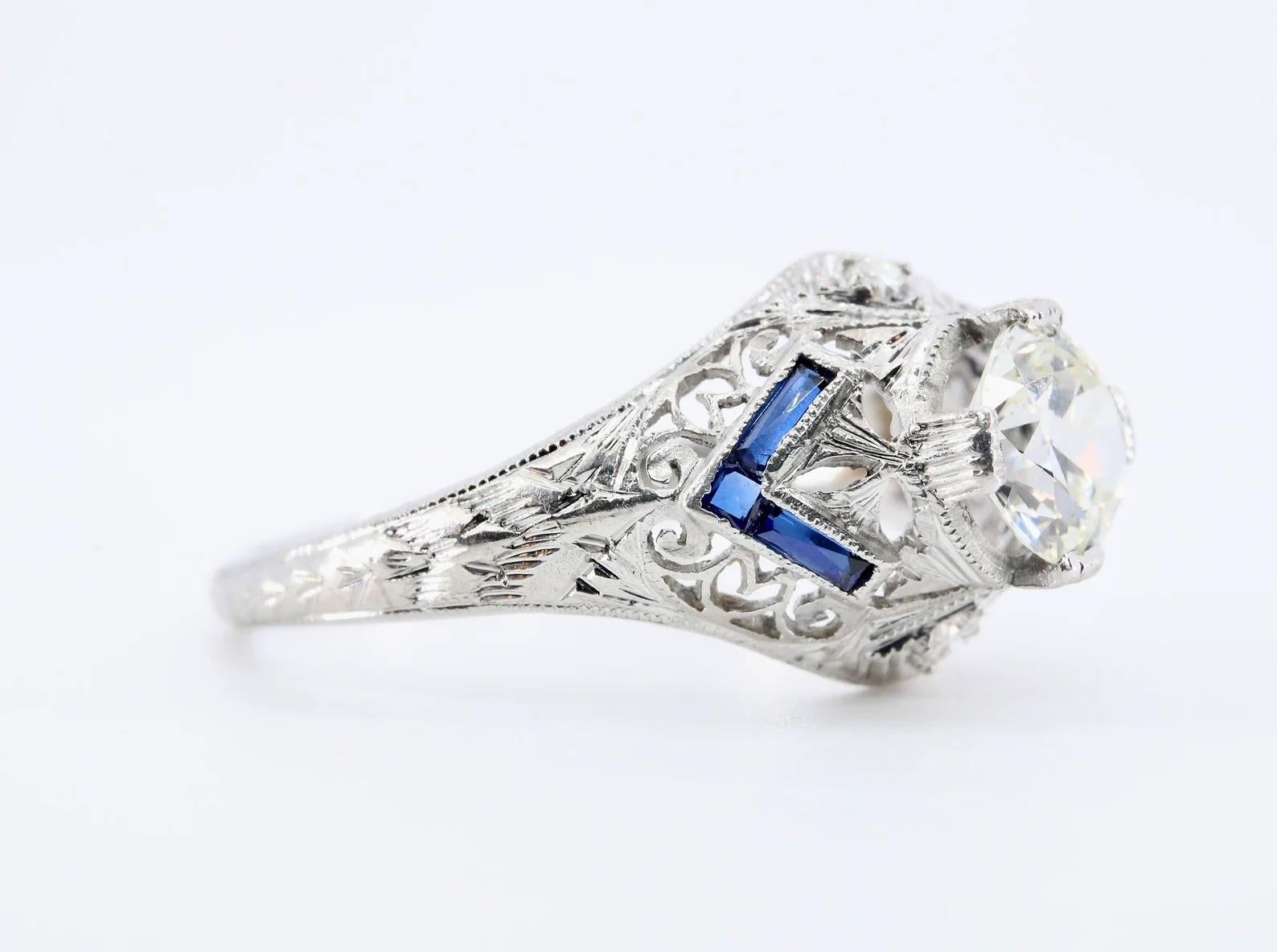 A handmade Art Deco period diamond, and sapphire engagement ring crafted in platinum. Centered by a 0.85 carat old European cut diamond of H color and VS1 clarity. 

The hand engraved mounting displays beautiful pierced filigree work throughout in a