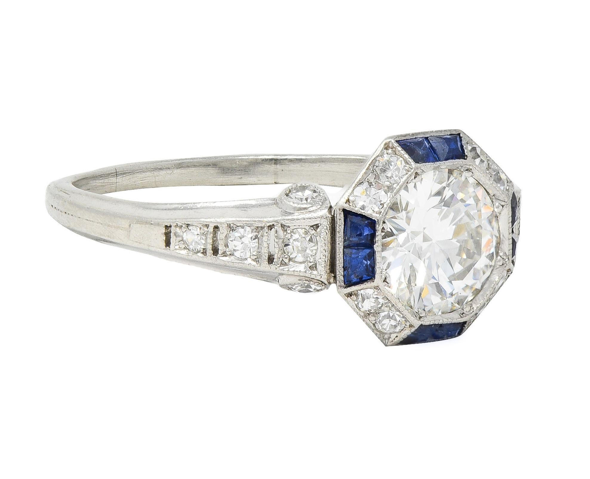 Centering an old European cut diamond weighing 0.90 carat total - G color with VS2 clarity 
Bead set flush in an octagonal form head with a faceted halo surround 
Featuring calibré cut sapphires weighing approximately 0.28 carat total
Transparent