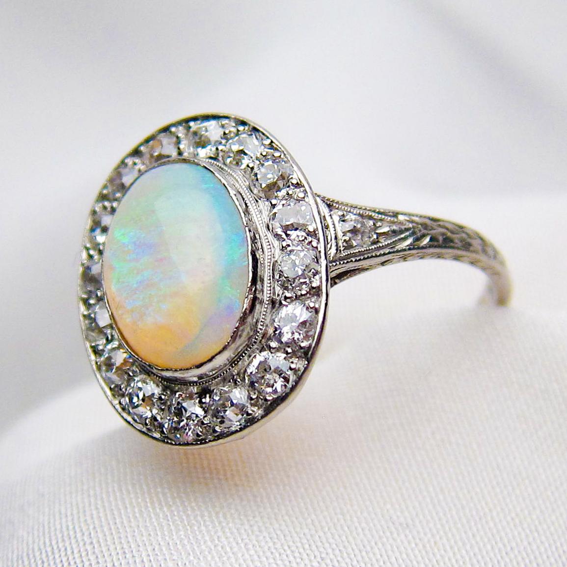 Circa 1920. This lovely Art Deco ring features a cabochon-cut 1.40 carat opal bezel-set in platinum. The opal features a blue and green play of color with a broad flash color pattern and is surrounded by a halo of 16 round old European-cut diamonds.