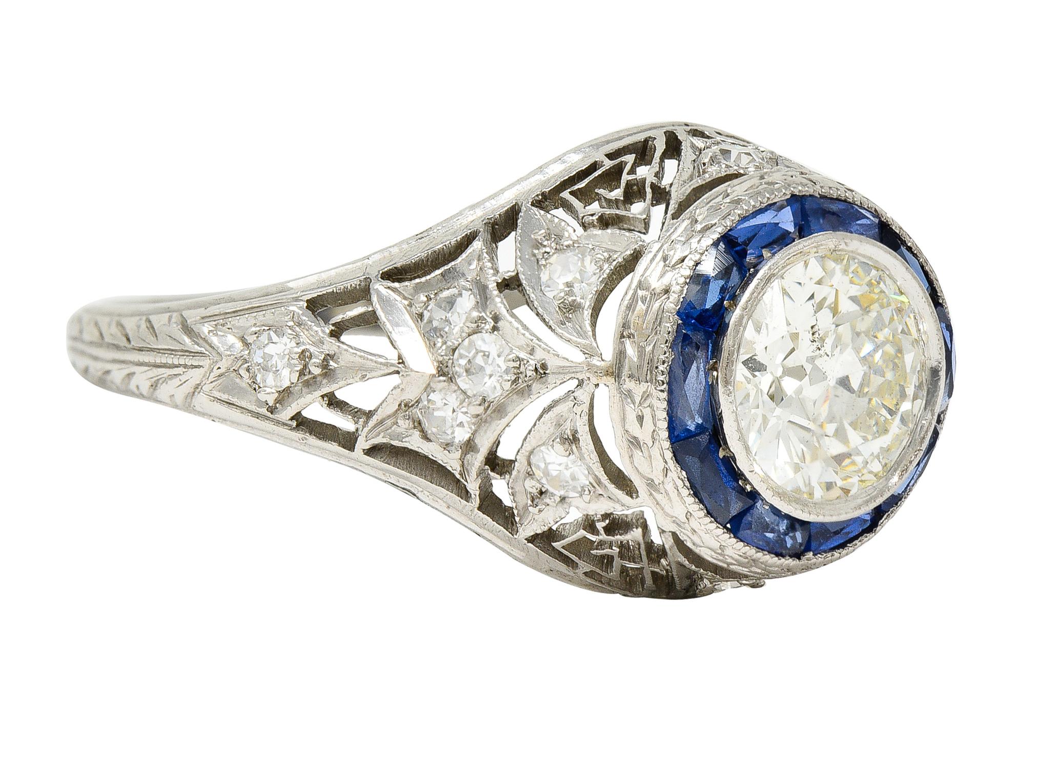 Centering an old European cut diamond weighing approximately 1.05 carats total - J color with SI2 clarity. Bezel set with a recessed French cut synthetic sapphire halo surround - channel set. Transparent medium blue in color - with an engraved wheat