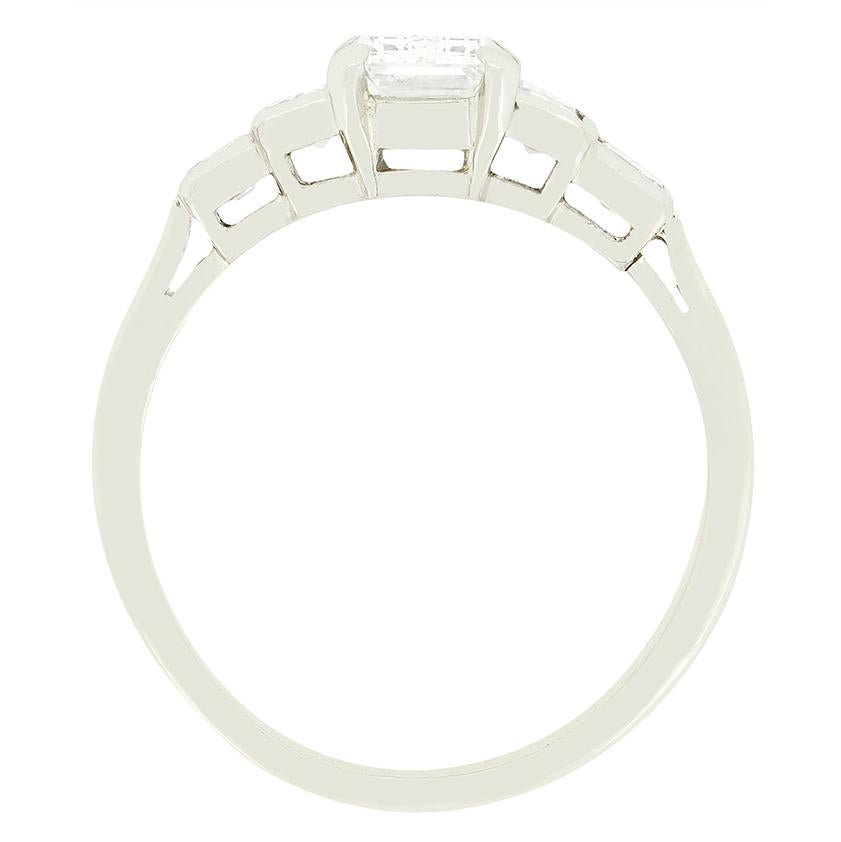 Taking centre stage in this beautiful Art Deco solitaire ring is a 1.50 carat, emerald cut diamond. The diamond has been estimated as I in colour and VS1 in clarity. Flanking the show piece stone, a pair of baguette cut diamonds are sat with two