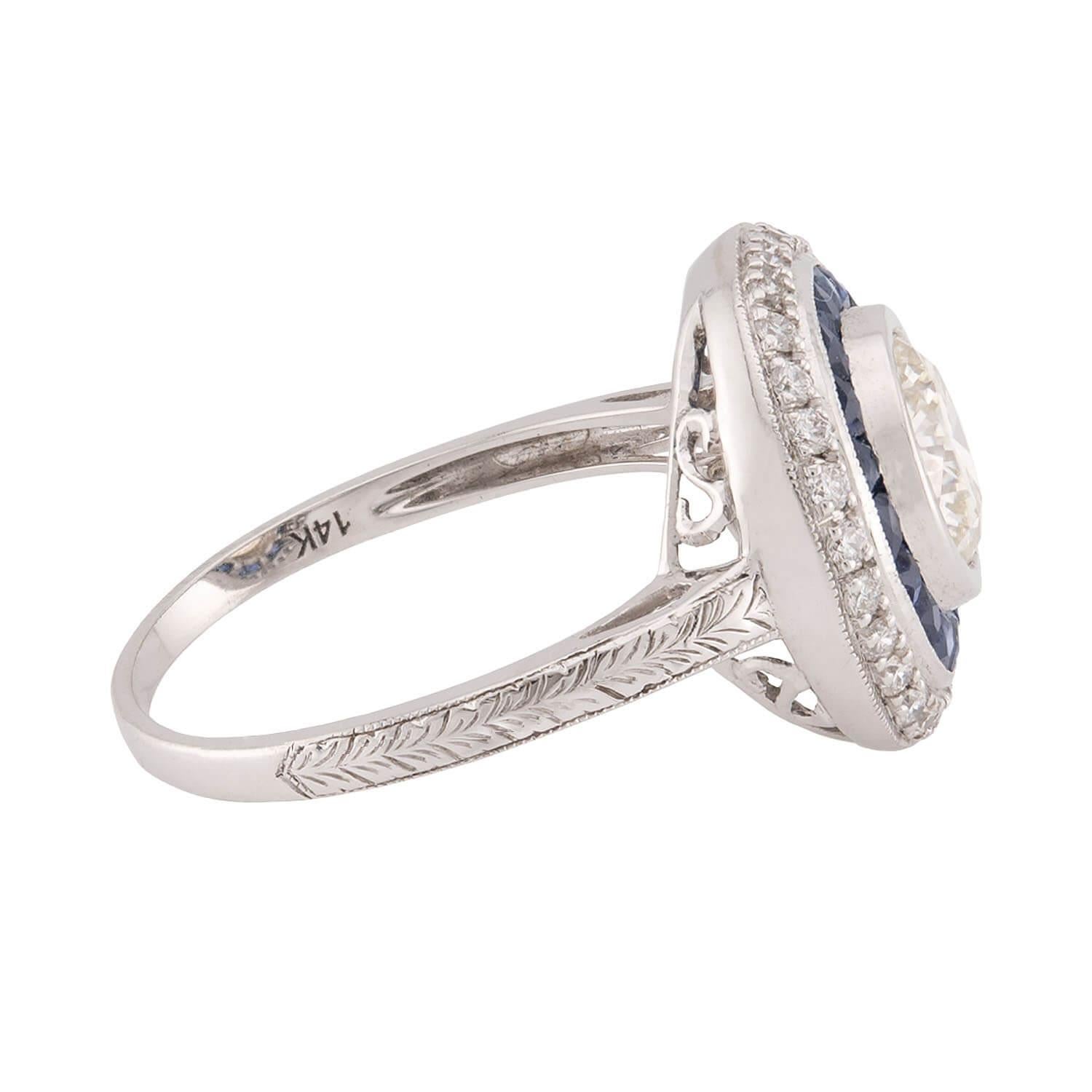 A truly magnificent diamond and sapphire halo ring from the Art Deco (ca1930s) era! Crafted in 14k white gold, this ring features a central .97ct old European cut diamond. The stone is held within a smooth and simple bezel-setting, and displays L/M