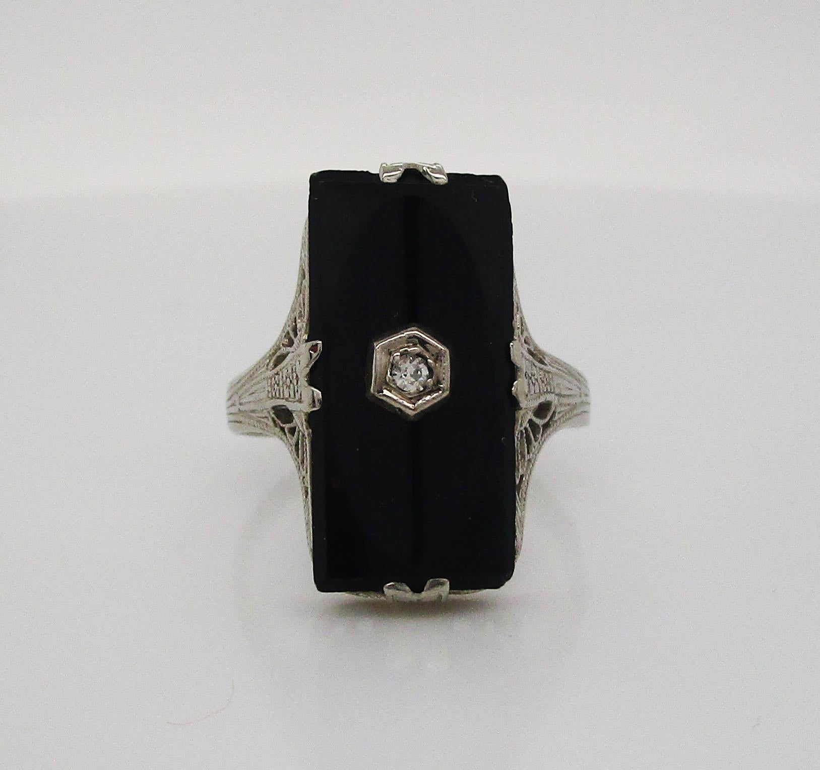 This absolutely beautiful Art Deco ring is in 14k white gold with delicate filigree details framing a black onyx center set with a stunning white diamond. The long north-to-south layout of the ring displays the dramatic black onyx rectangle that