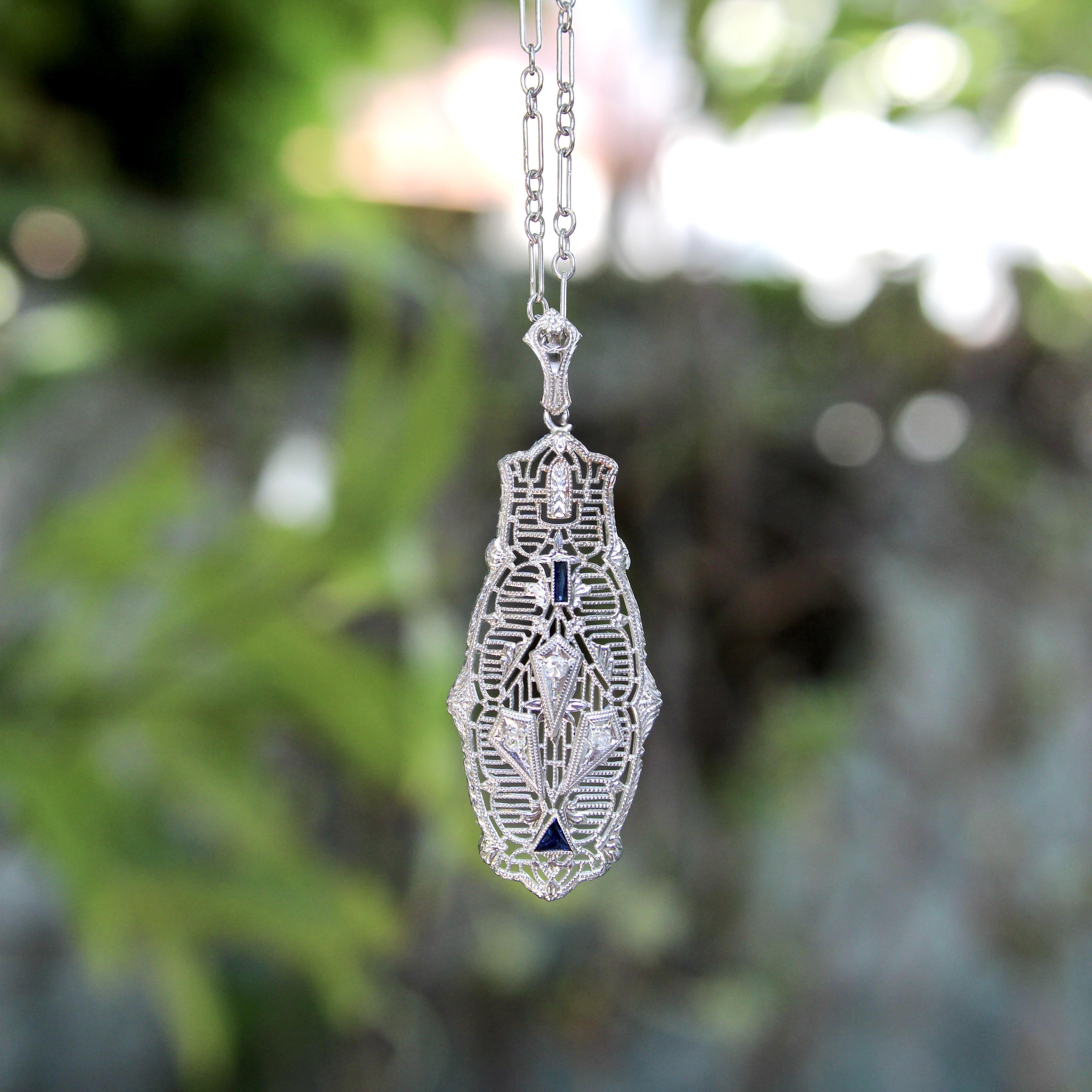 Circa the cusp of the Edwardian and Art Deco eras (1915-1920), this 14k white gold pendant necklace has beautiful filigree work that is open and lace like. Within the filigree pattern, there are horizontal lines along the lozenge part of the