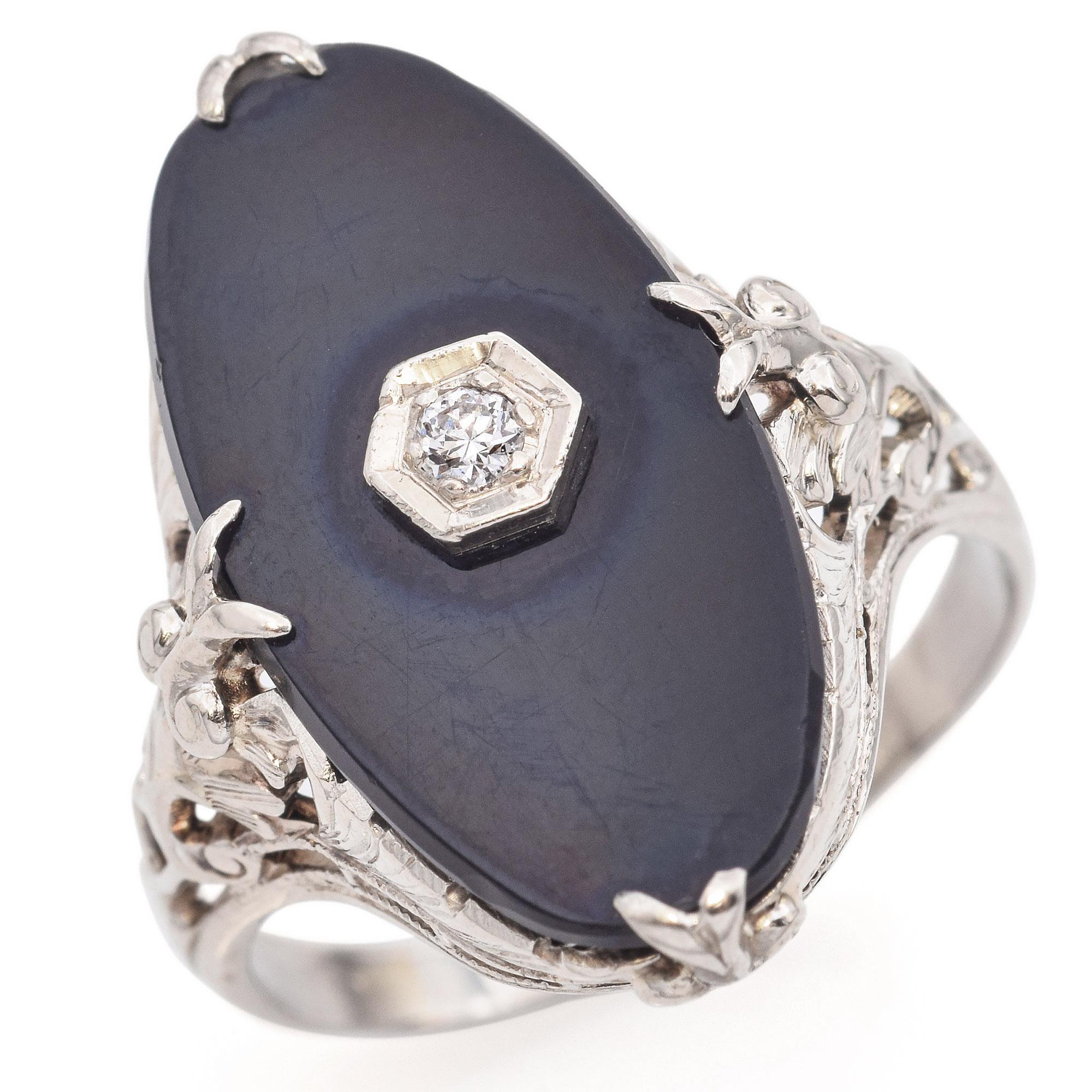 This exquisite ring features a striking black onyx centerpiece, elegantly set in 14k white gold. The onyx is adorned with a single sparkling diamond, adding a touch of brilliance. The intricate filigree detailing on the band enhances its