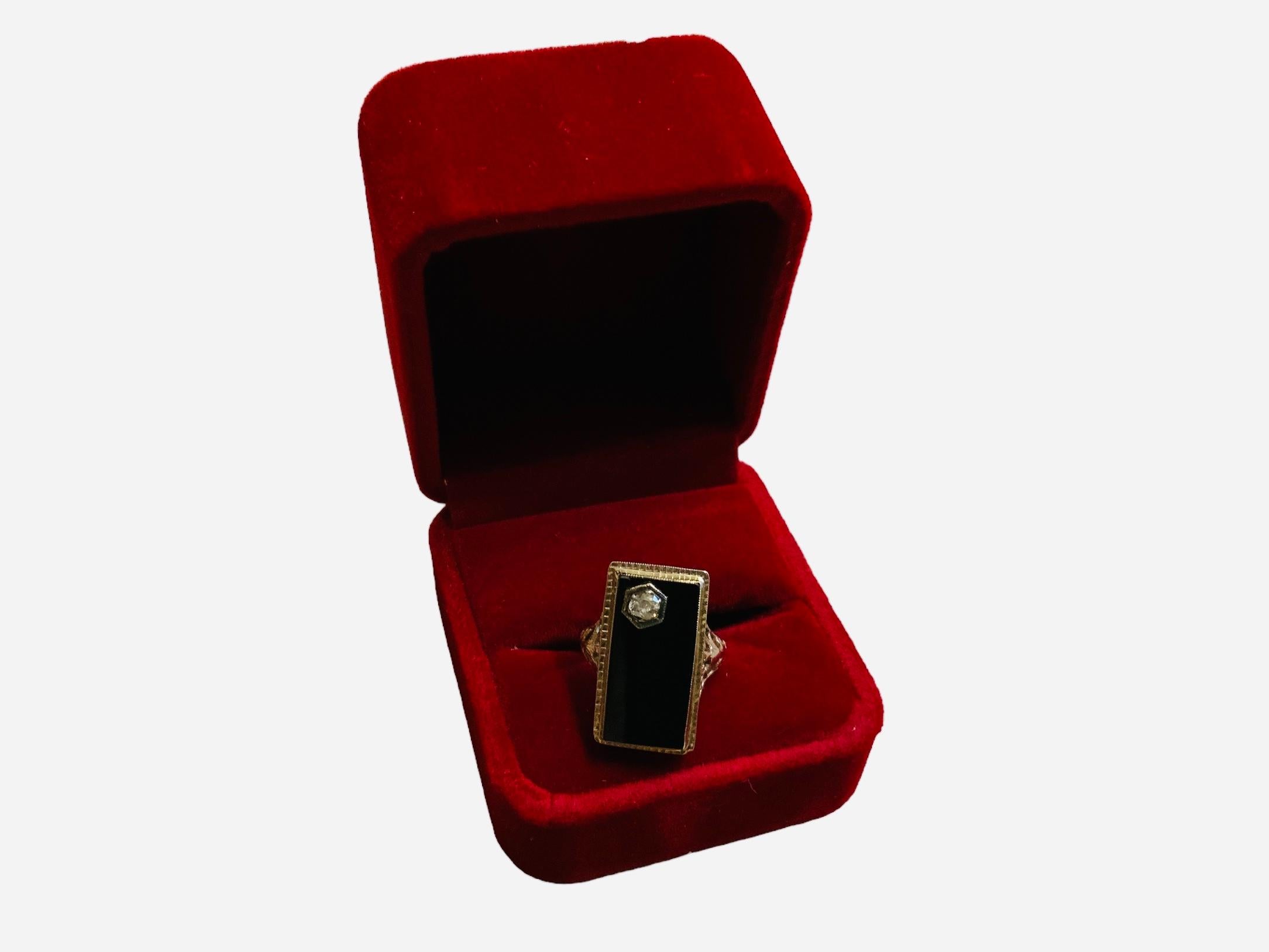 This is an Art Deco 14K white gold, diamond and onyx cocktail ring. It depicts a rectangular black onyx top in white gold bezel setting. The onyx has a reeded gold frame. A round cut diamond on prong setting embellishes one of the corner of the