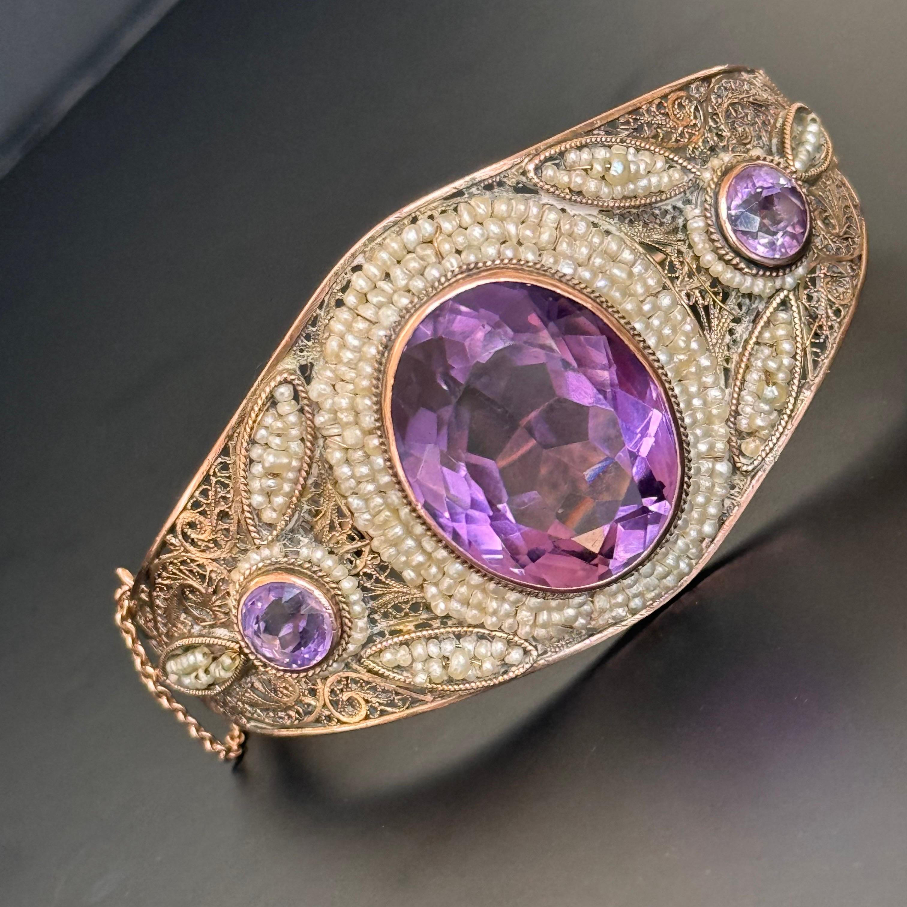 This stunning Art Deco hinged bangle bracelet is a true masterpiece of early 20th-century craftsmanship. Set in 14kt gold, the bracelet features a large central amethyst stone, measuring 7/8 inches x 3/4 inches x 3/8 inches in depth, which is