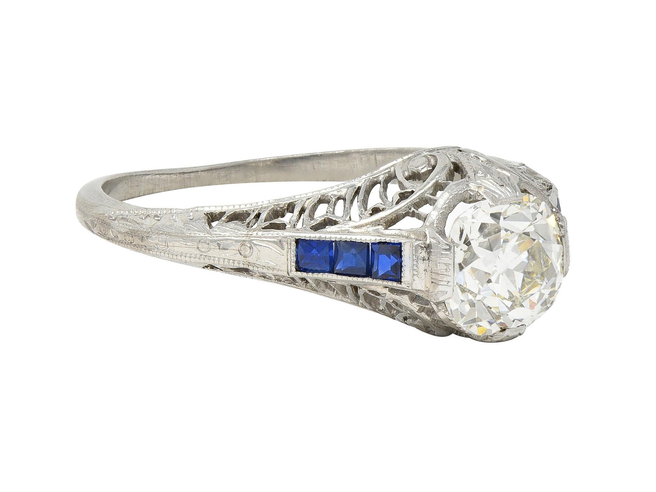 Centering an old mine cut diamond weighing approximately 1.28 carats - K color with VS2 clarity
Set with engraved wide prongs with a pierced scrolling plume motif gallery 
Flanked by rows of step cut sapphires channel set in shoulders
Weighing