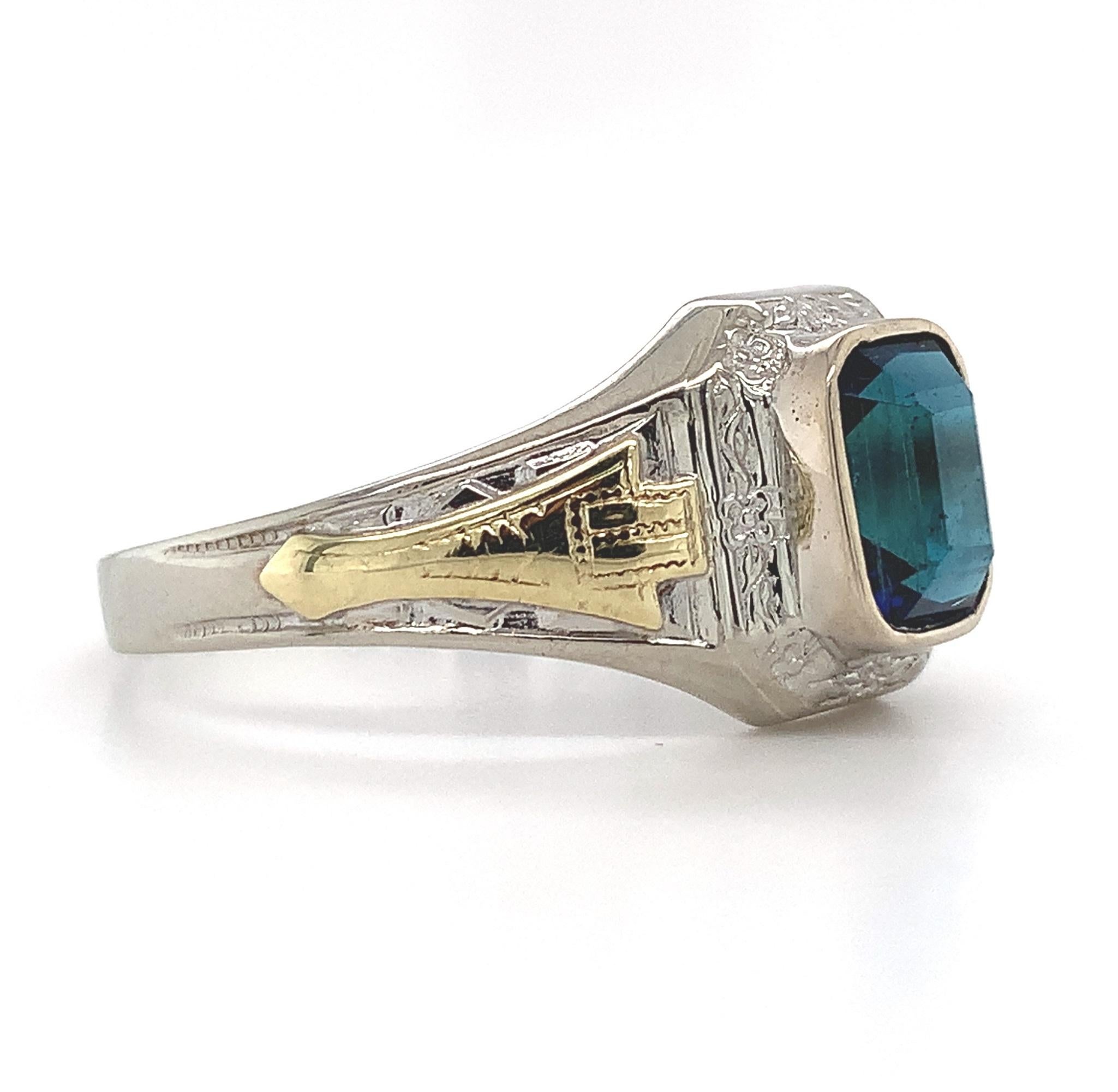 10K white gold Art Deco ring with applied yellow gold accents. The ring features an emerald cut blue-green tea color tourmaline weighing 1.54 carats. The tourmaline measures about 704mm x 5.7mm and has a small chip. There is a new bezel. The ring